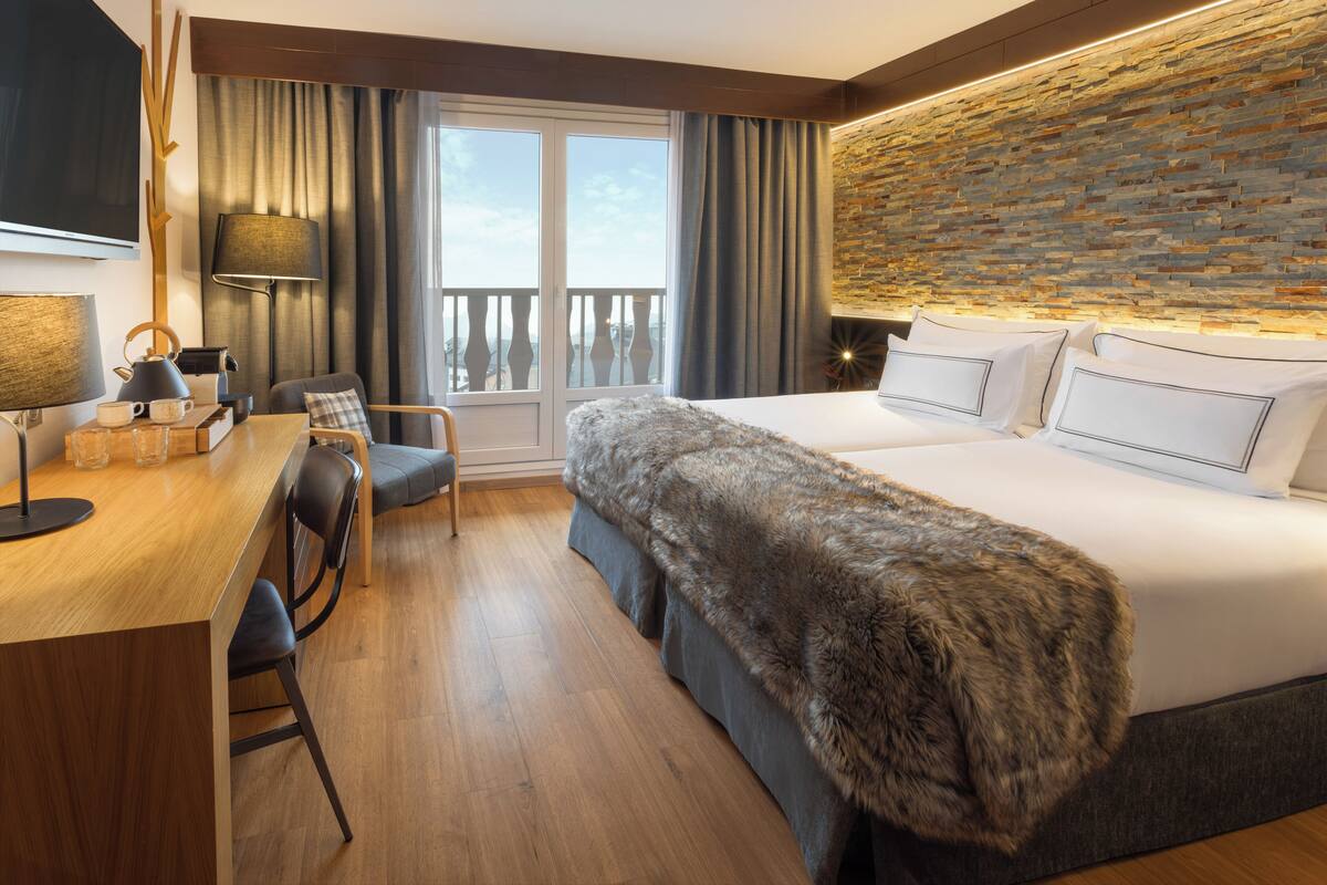 A modern hotel room with a large bed, a desk with a chair, a television, and a balcony with curtains.