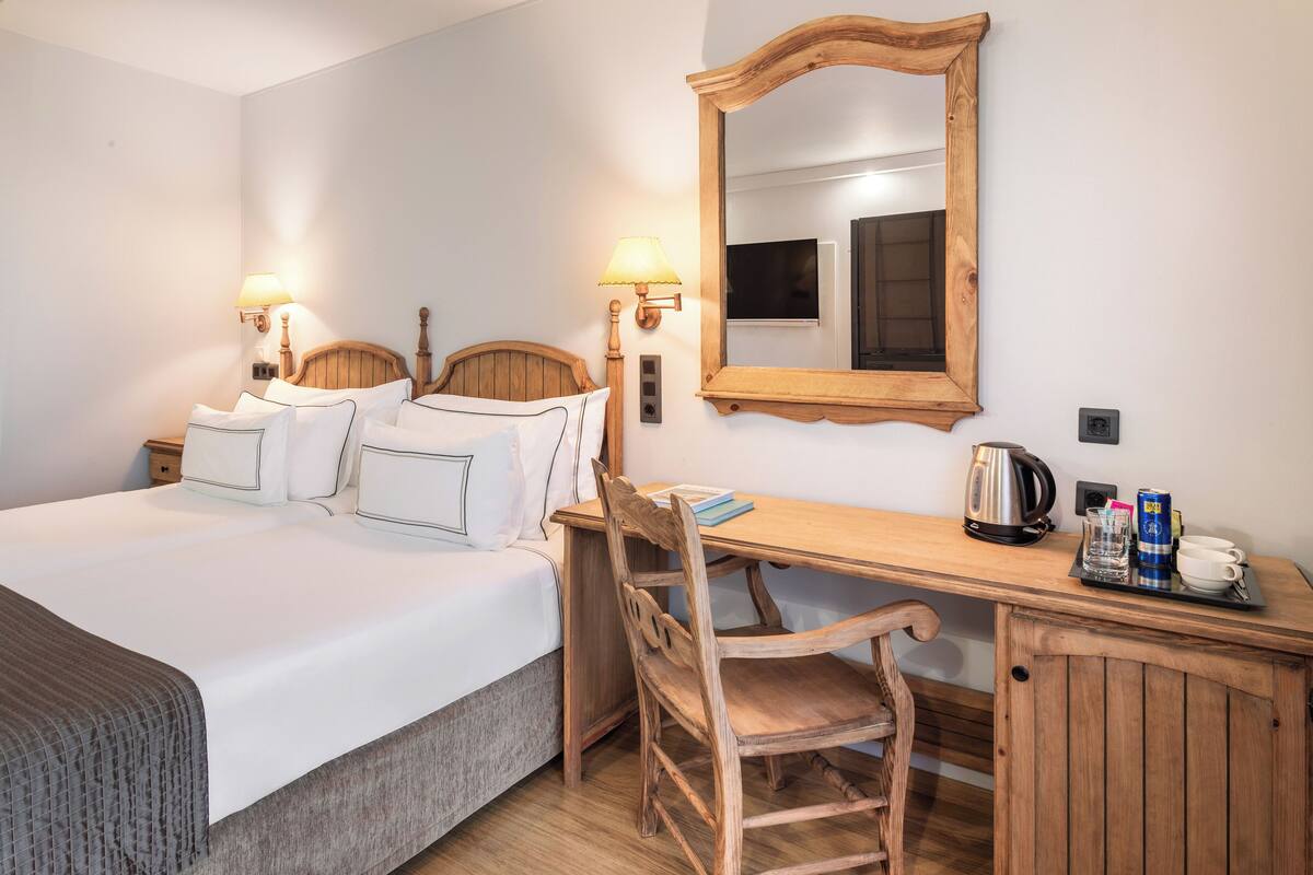 A hotel room with a bed, bedside lamps, a wooden desk, a chair, a mirror, and a tray with a coffee pot, cups, and a can.