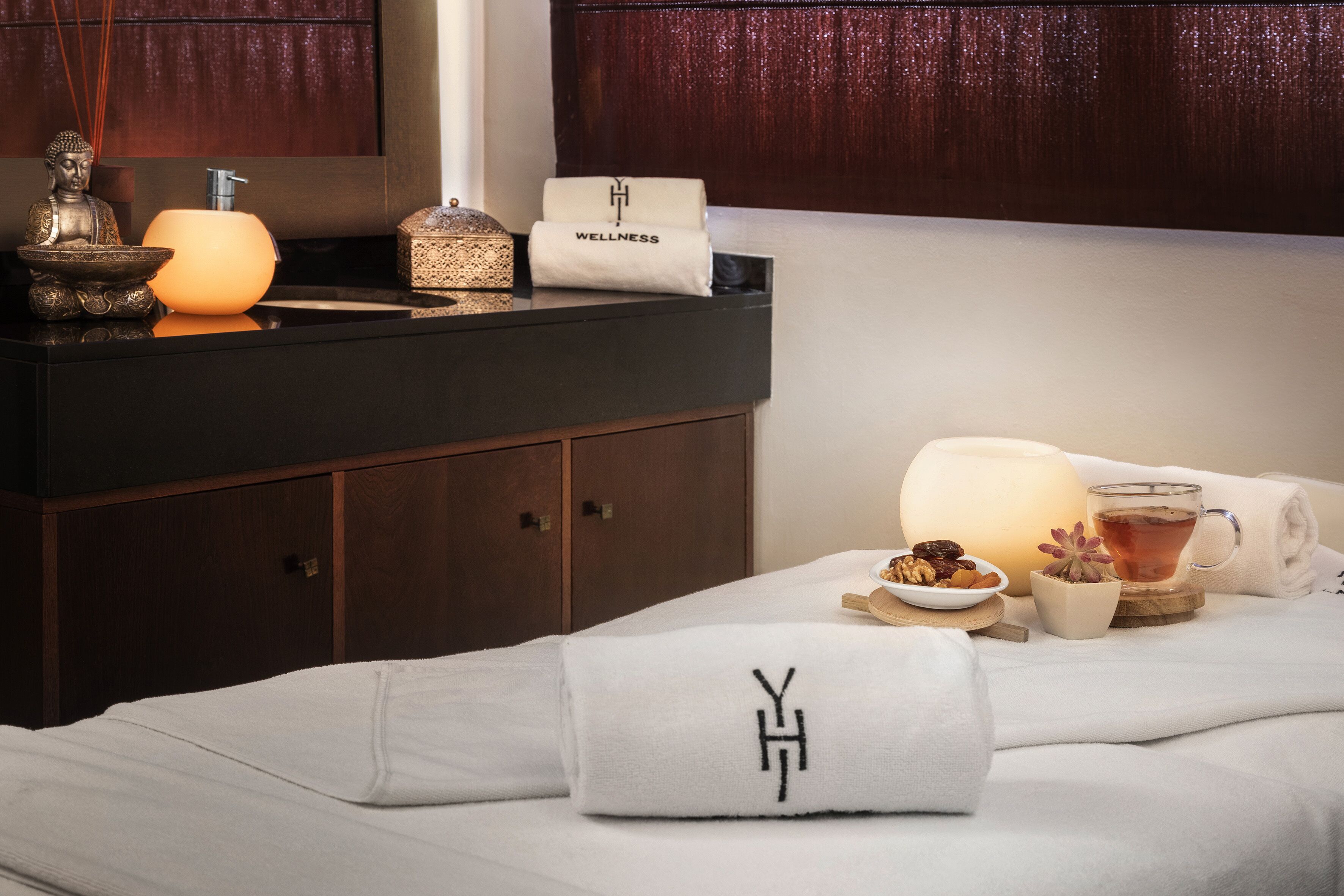 A neatly arranged hotel room with a bed, a bedside table with a lamp, a Buddha statue, and a wellness-themed towel.
