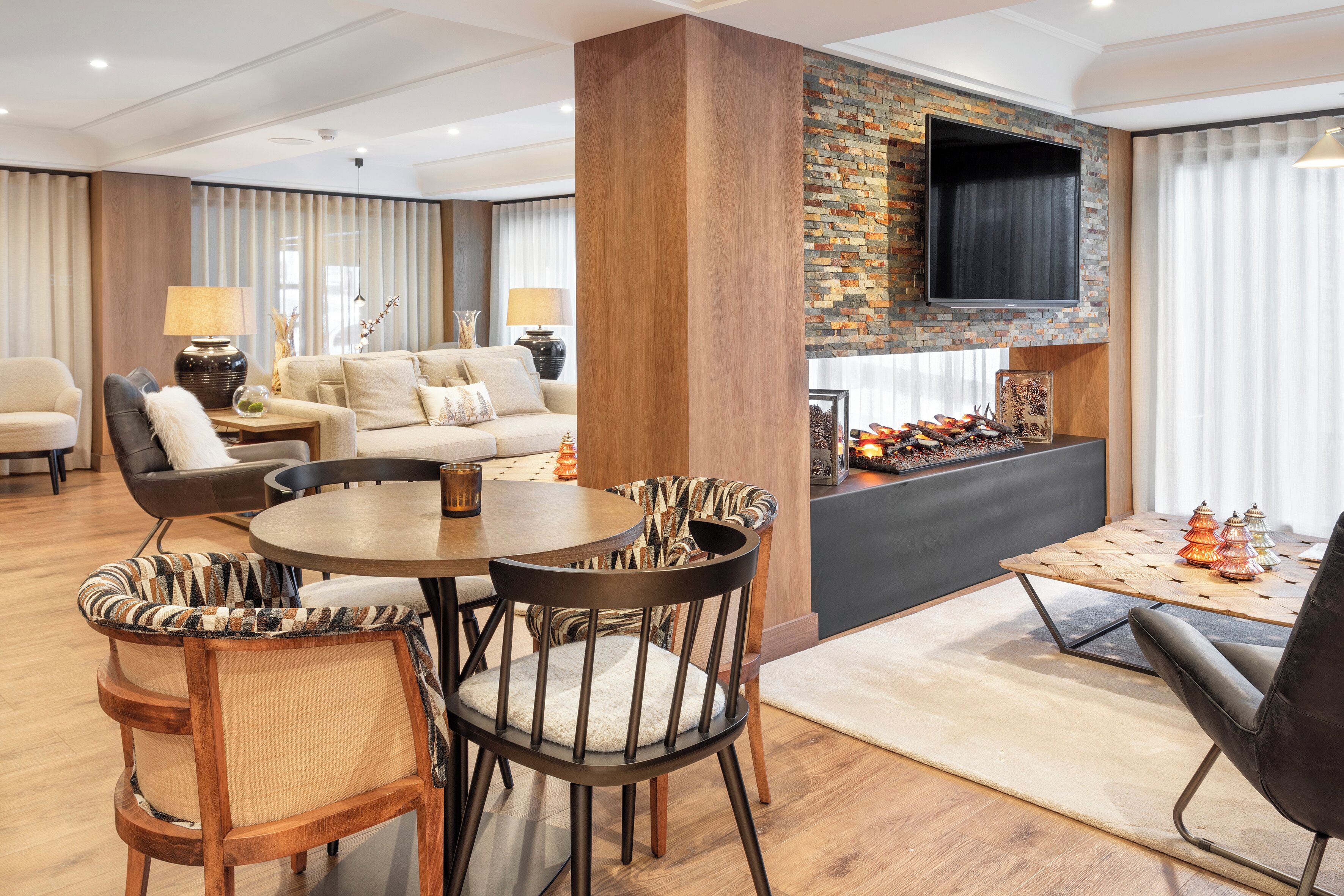 A modern living room with a round dining table, wooden chairs, a sofa, and a flat-screen TV mounted on a brick wall.