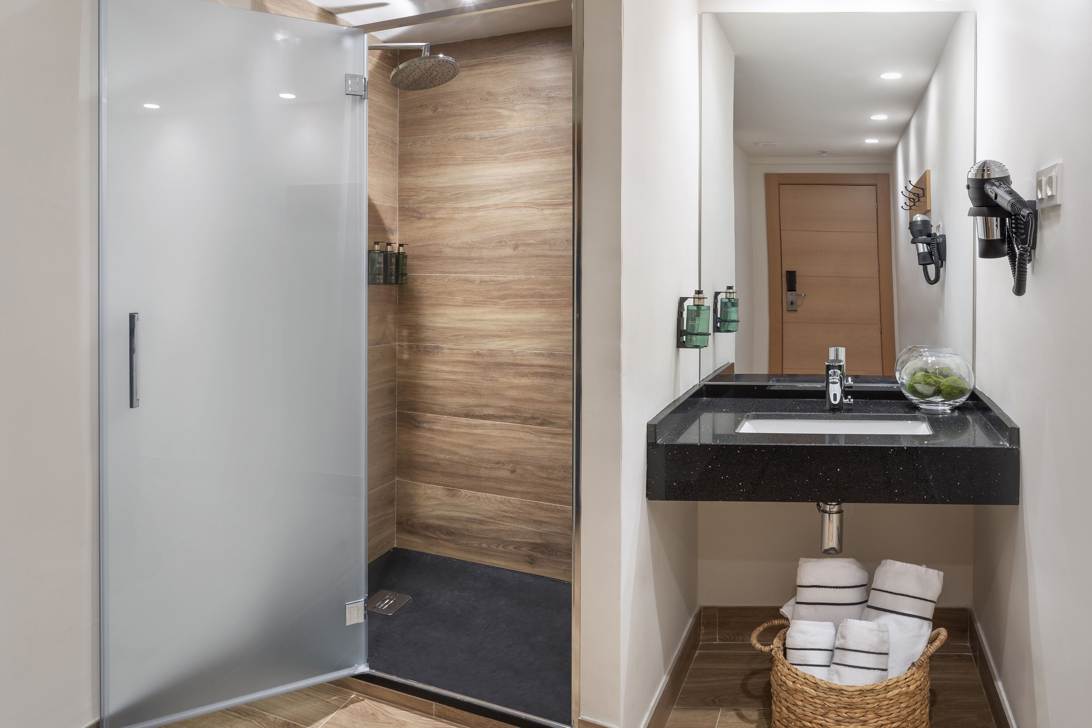 A modern bathroom with a glass-enclosed shower, a black countertop sink, and a mirror with a hairdryer mounted on the wall.