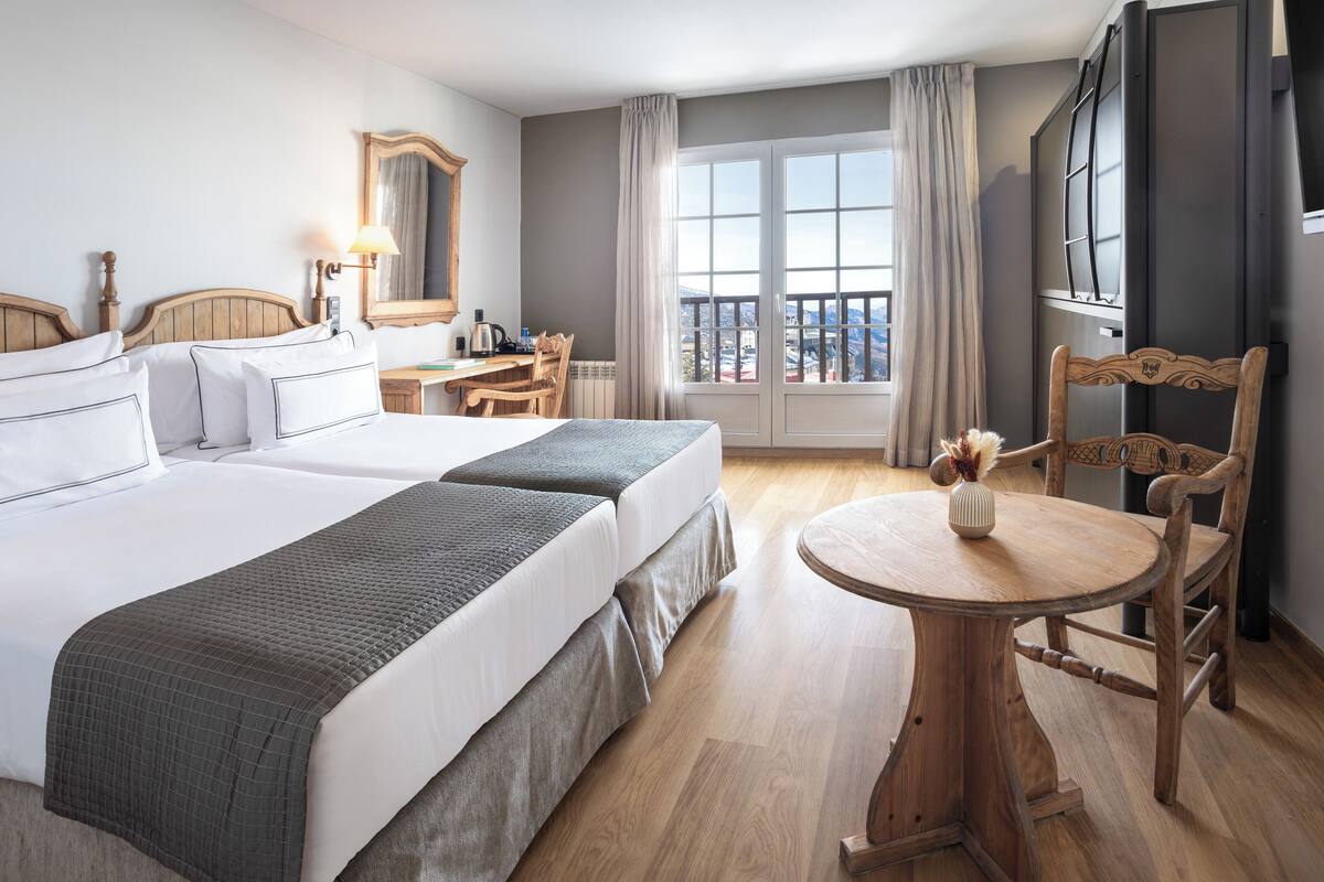 A hotel room with two beds, a desk, a chair, a round wooden table, and a large window with curtains.