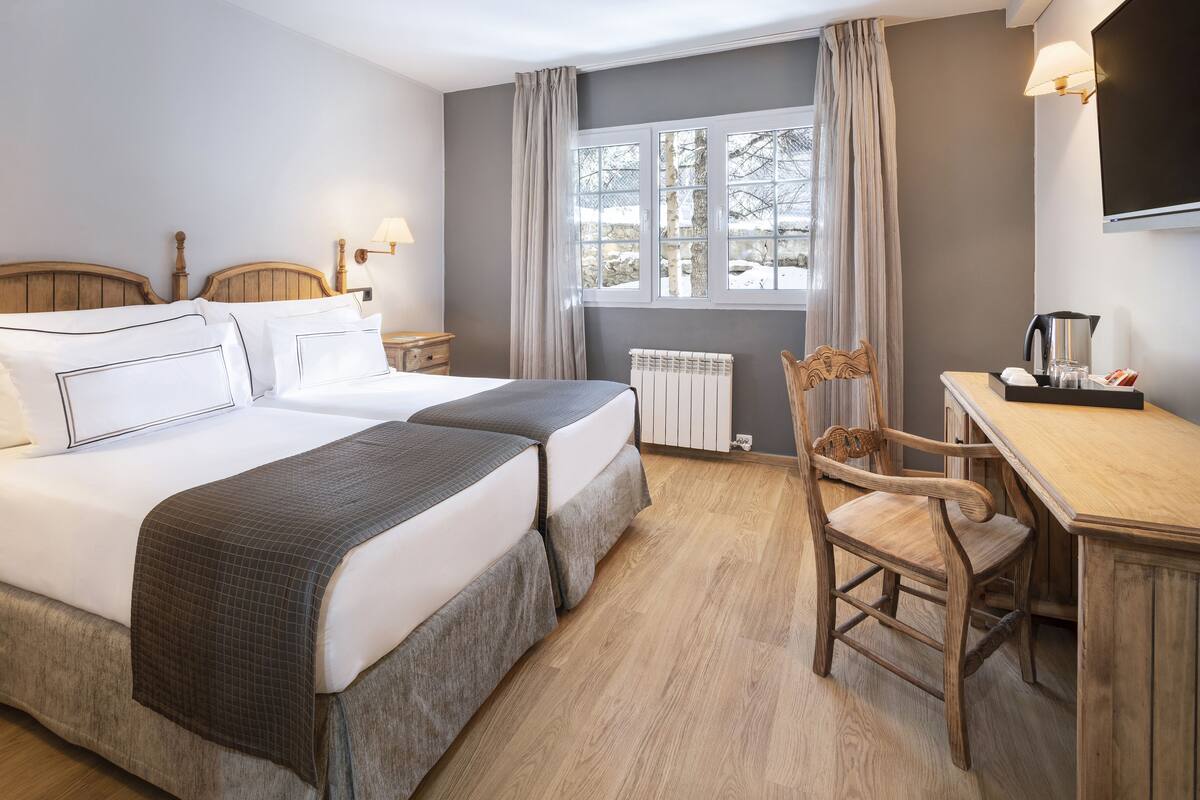 A hotel room with two beds, a wooden desk, a chair, a radiator, and a window with curtains.
