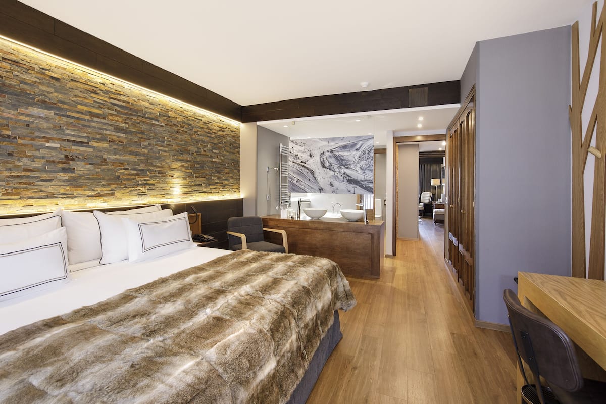 A modern hotel room with a large bed, a wooden desk, a bathroom with a marble wall, and a hallway leading to another room.