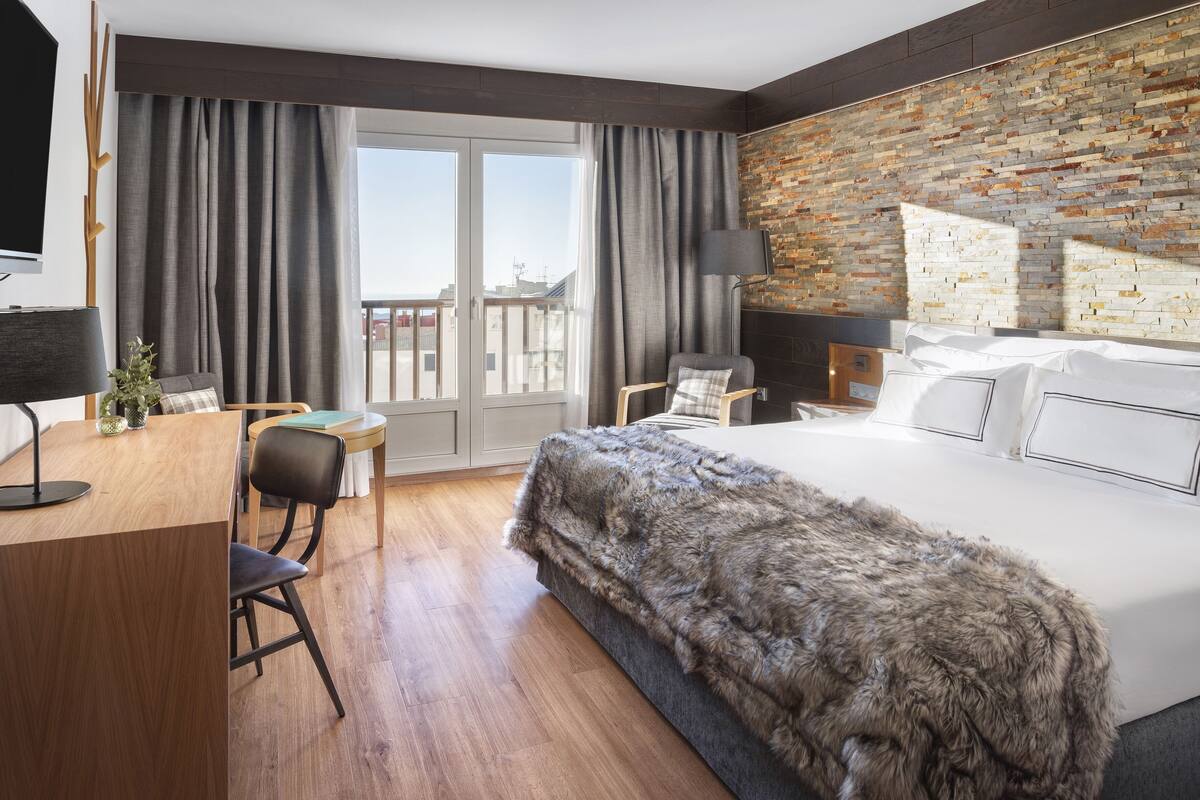 A modern hotel room with a large bed, a desk with a chair, a small table, and a balcony with curtains.