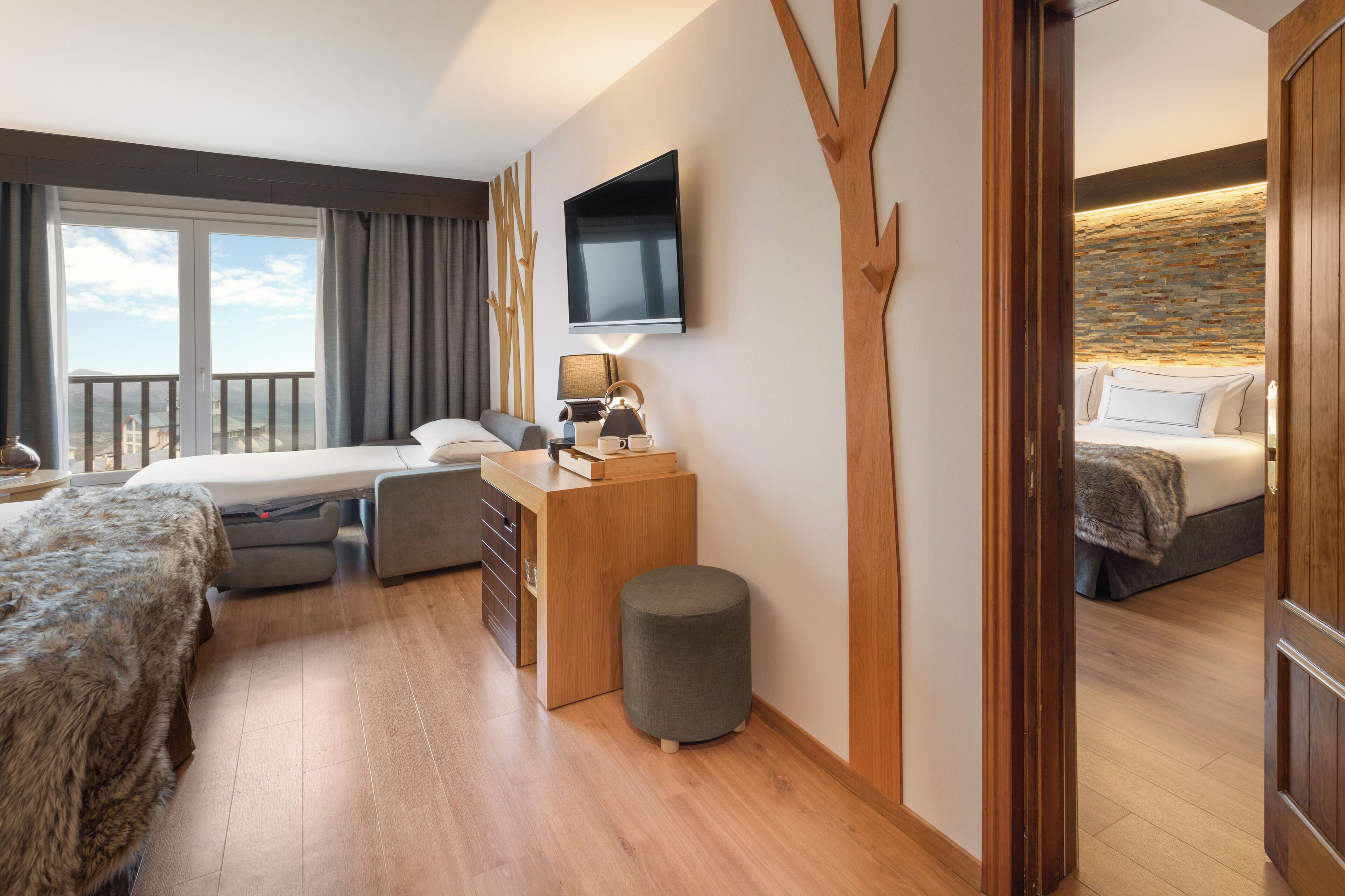 A modern hotel room with a large bed, a desk, a chair, a small ottoman, a TV, and a balcony with a view.