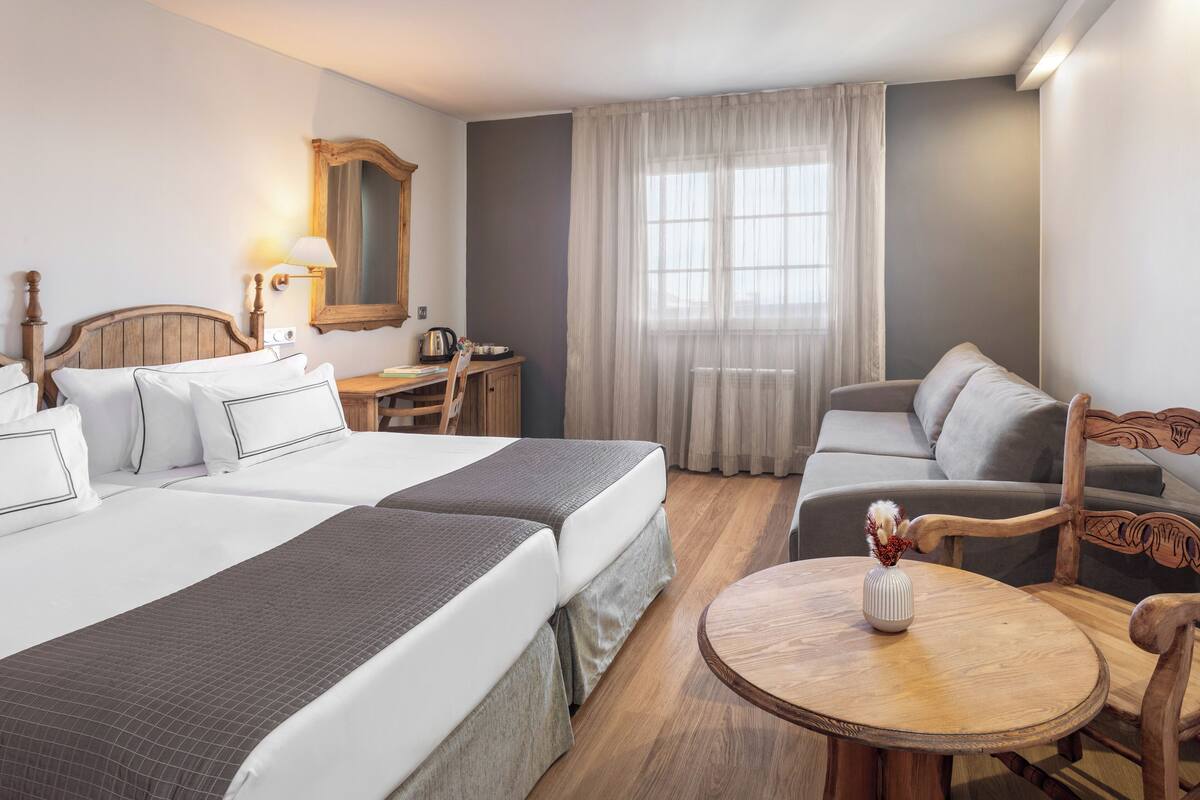 A hotel room with two beds, a desk, a chair, a round wooden table, and a large window with curtains.