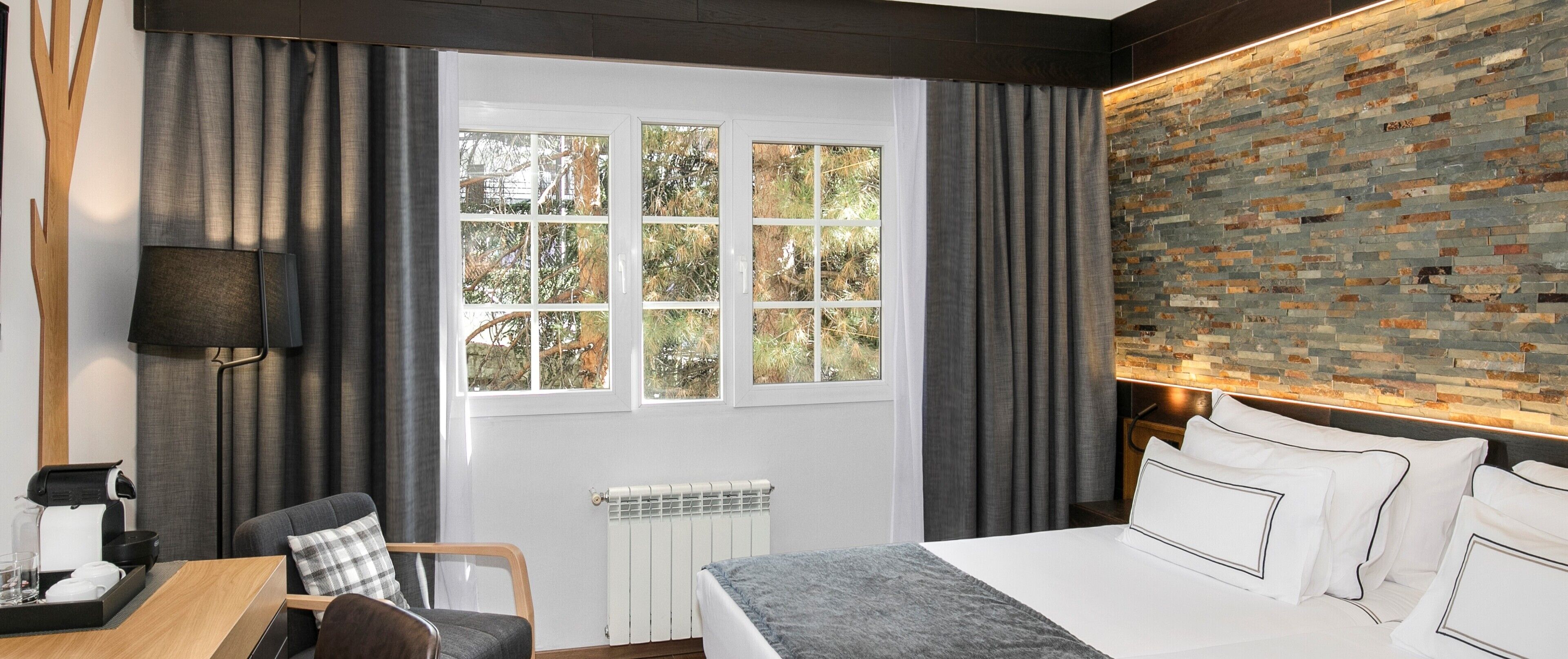 Habitación de hotel con un ventanal grande, pared con diseño de ladrillo, cama con ropa blanca, escritorio con lámpara y una silla.