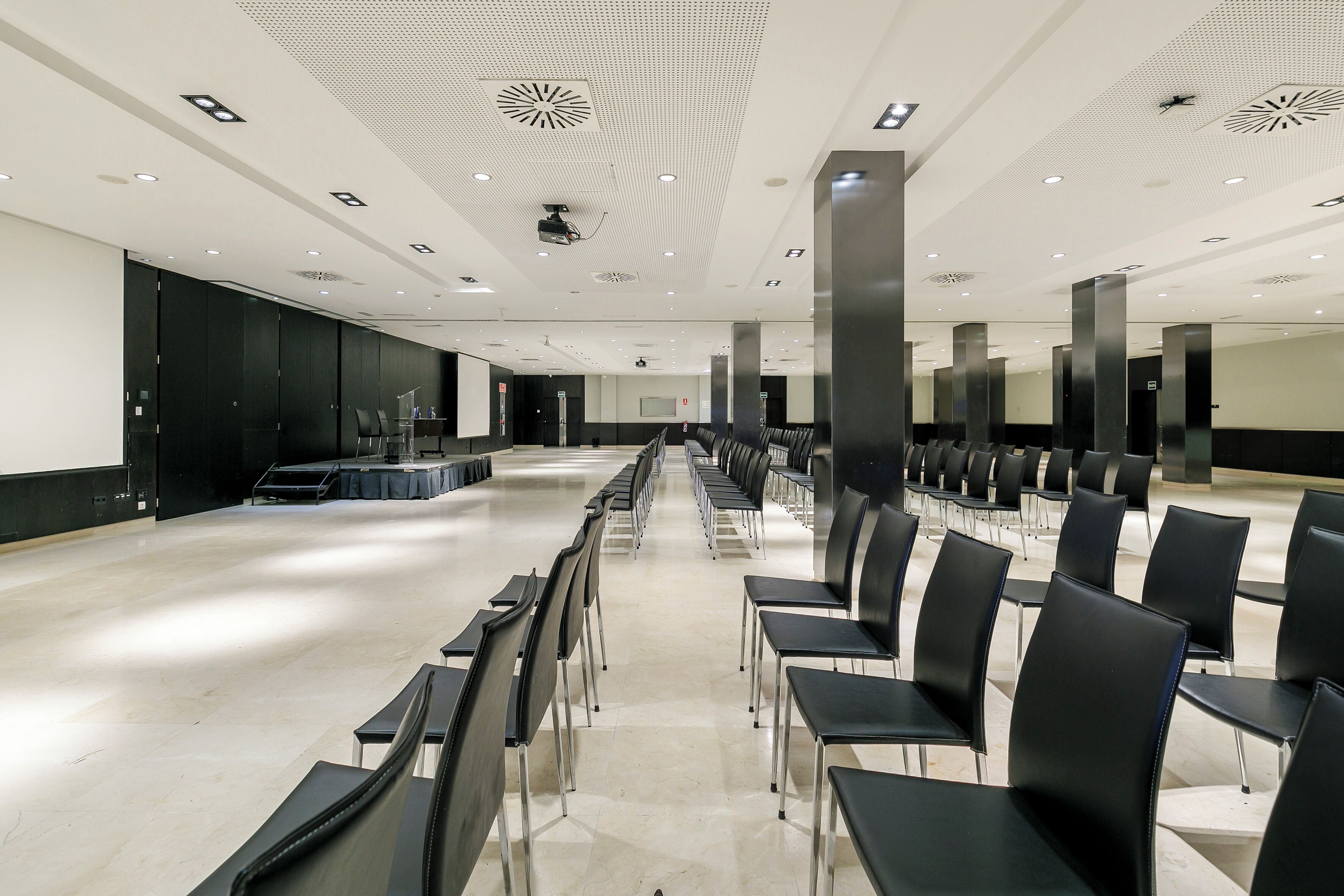 Ein geräumiger Konferenzraum mit Reihen schwarzer Stühle und einem langen Tisch, ausgestattet mit einem Projektor und einer Leinwand.