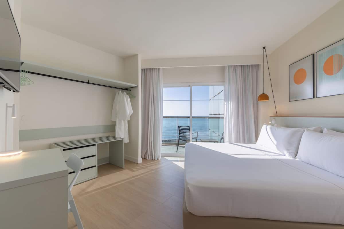 Habitación de hotel con cama, escritorio, silla, balcón con vista al mar y obras de arte en la pared.