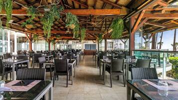 Un comedor interior con mesas y sillas, plantas colgantes y ventanas amplias.