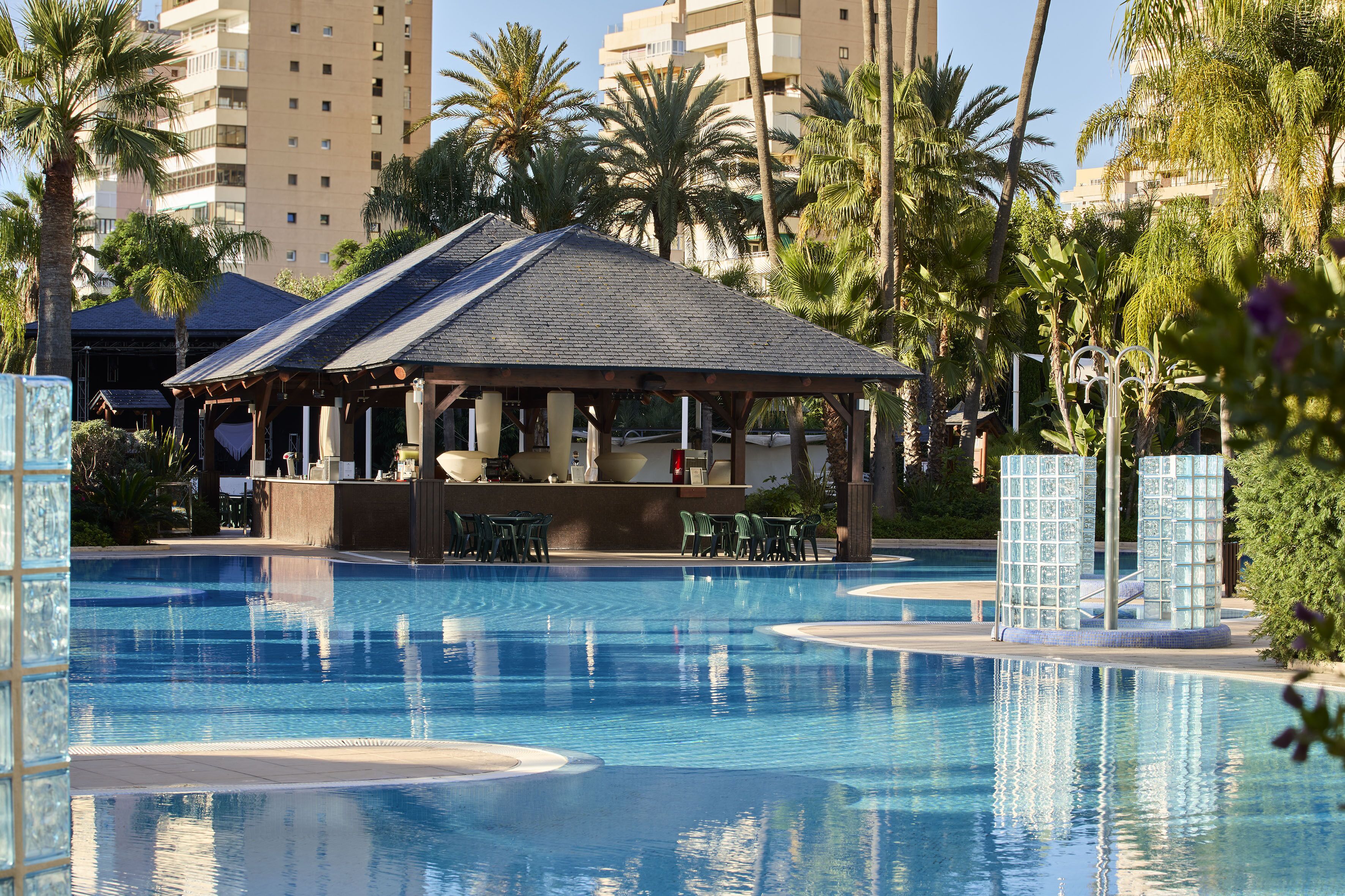 Una piscina con zona de estar cubierta, rodeada de palmeras y un edificio de varios pisos al fondo.