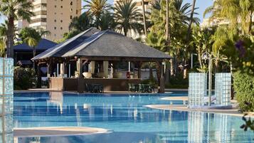 Una piscina con zona de estar cubierta, rodeada de palmeras y un edificio de varios pisos al fondo.