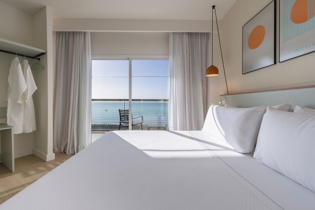 A hotel room with a large bed, a chair, a table, and a view of the sea.