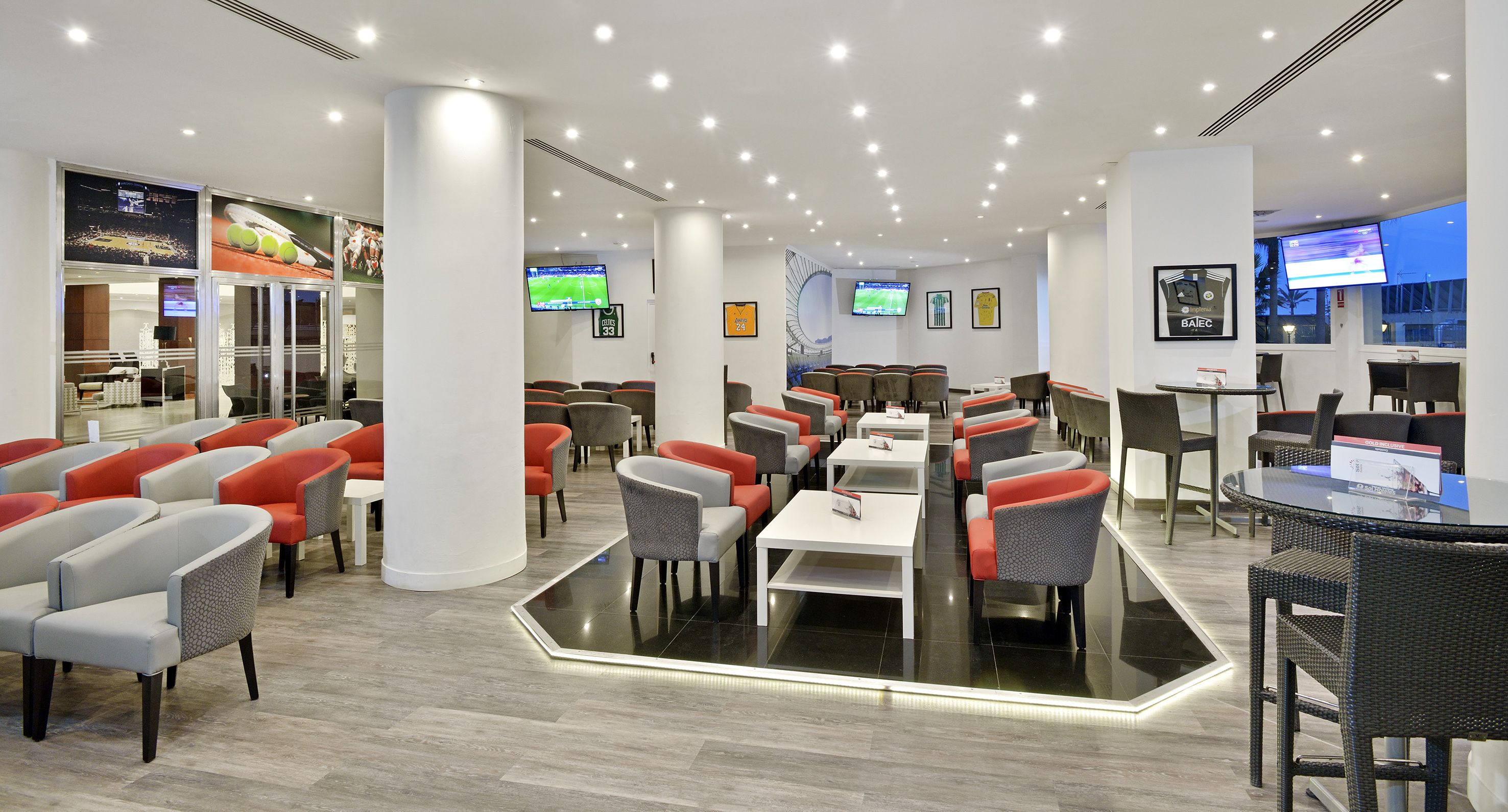 Eine moderne Hotelhalle mit mehreren Sitzbereichen, darunter ein zentraler Essbereich mit Tischen und Stühlen sowie eine Bar mit hohen Hockern.