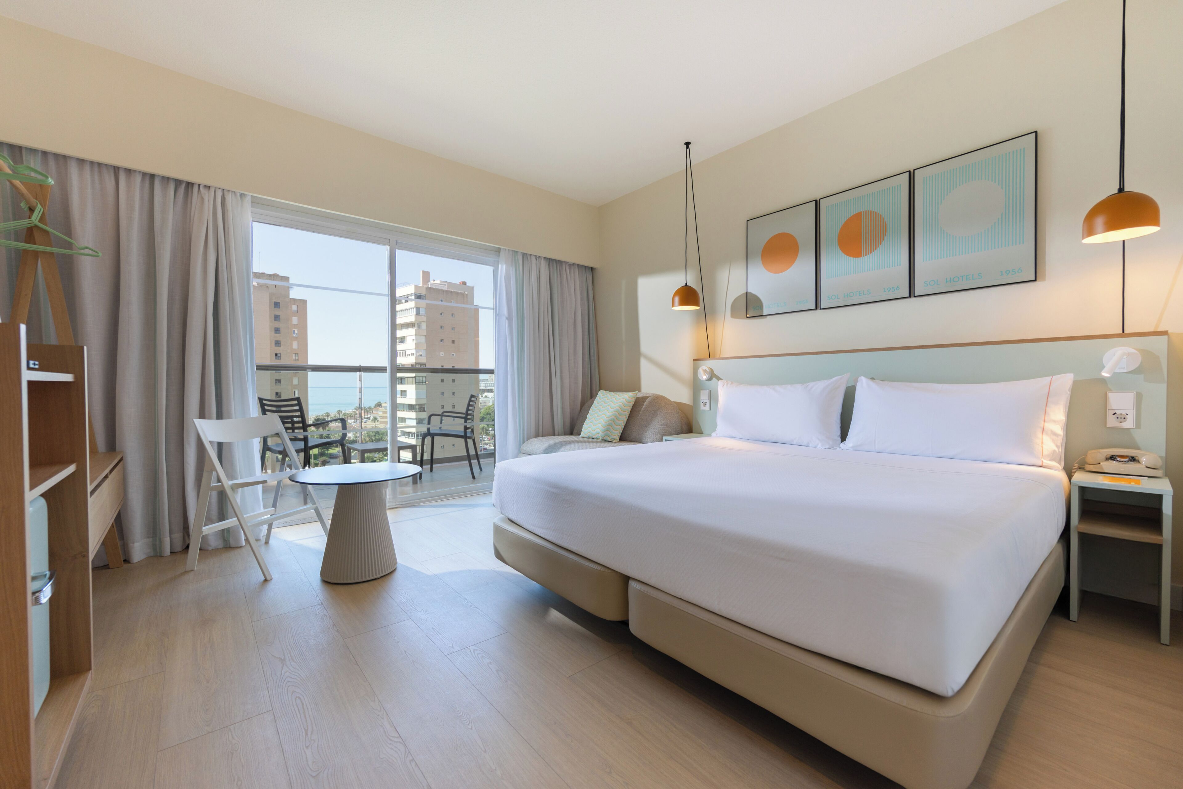 A modern hotel room with a large bed, a round dining table, a chair, a nightstand with a phone, and a view of the city through the window.