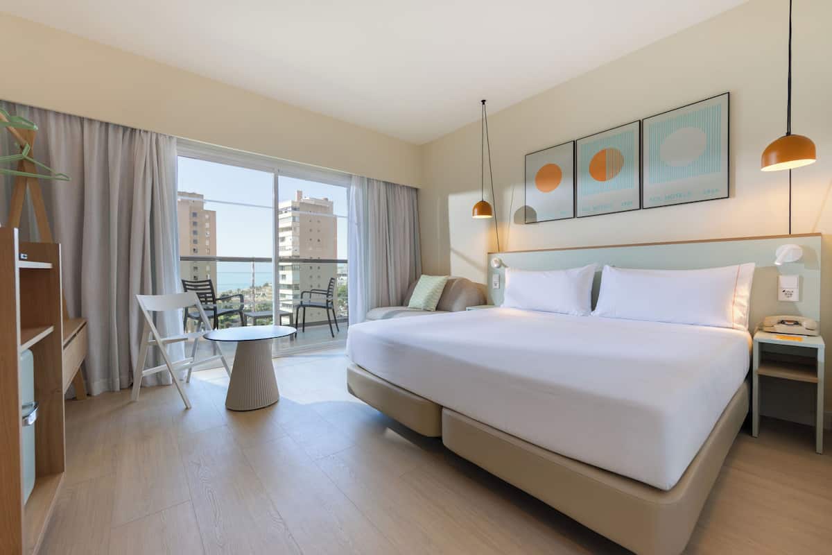 Una habitación de hotel moderna con una cama grande, una mesa de comedor redonda, una silla, una mesita de noche con un teléfono y vistas a la ciudad a través de la ventana.