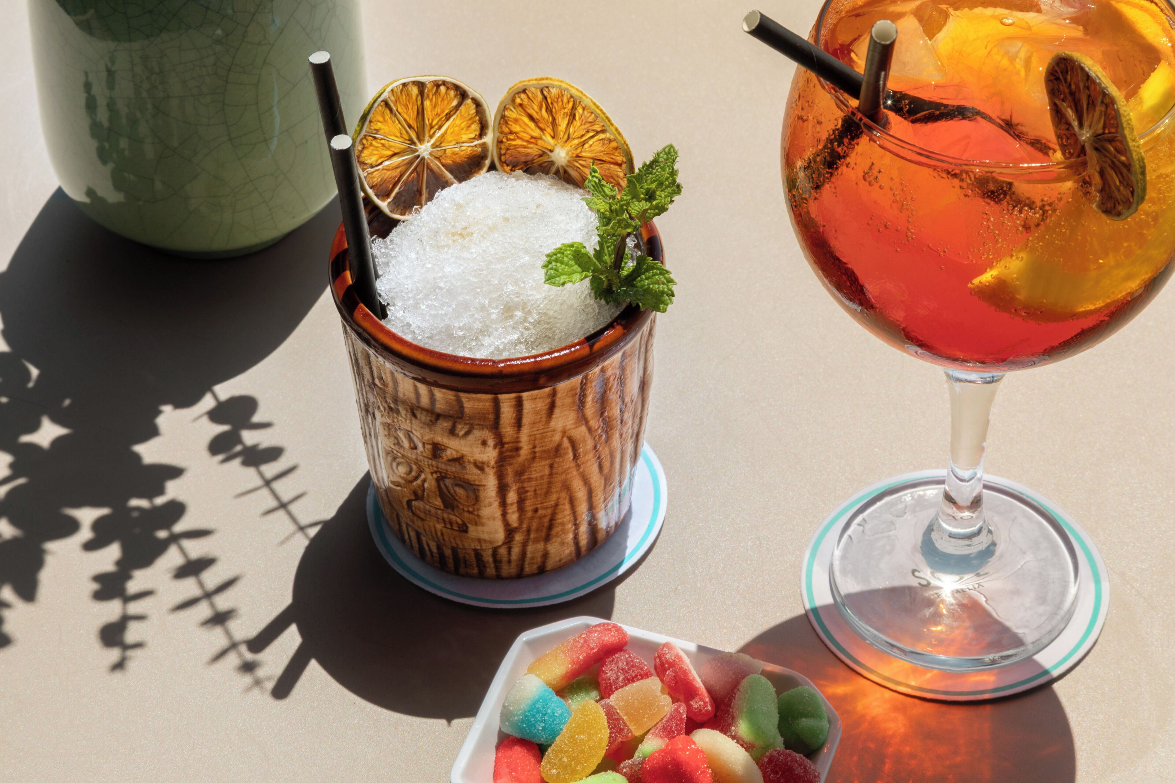 Two drinks, one in a wooden cup and one in a glass, with ice and garnishes.