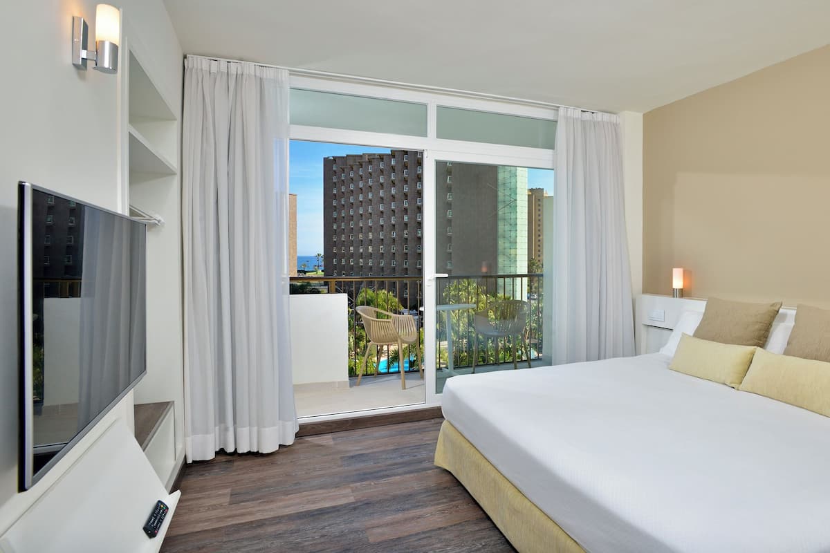 Habitación de hotel con cama, televisor, balcón y vistas a edificios, además de piscina.