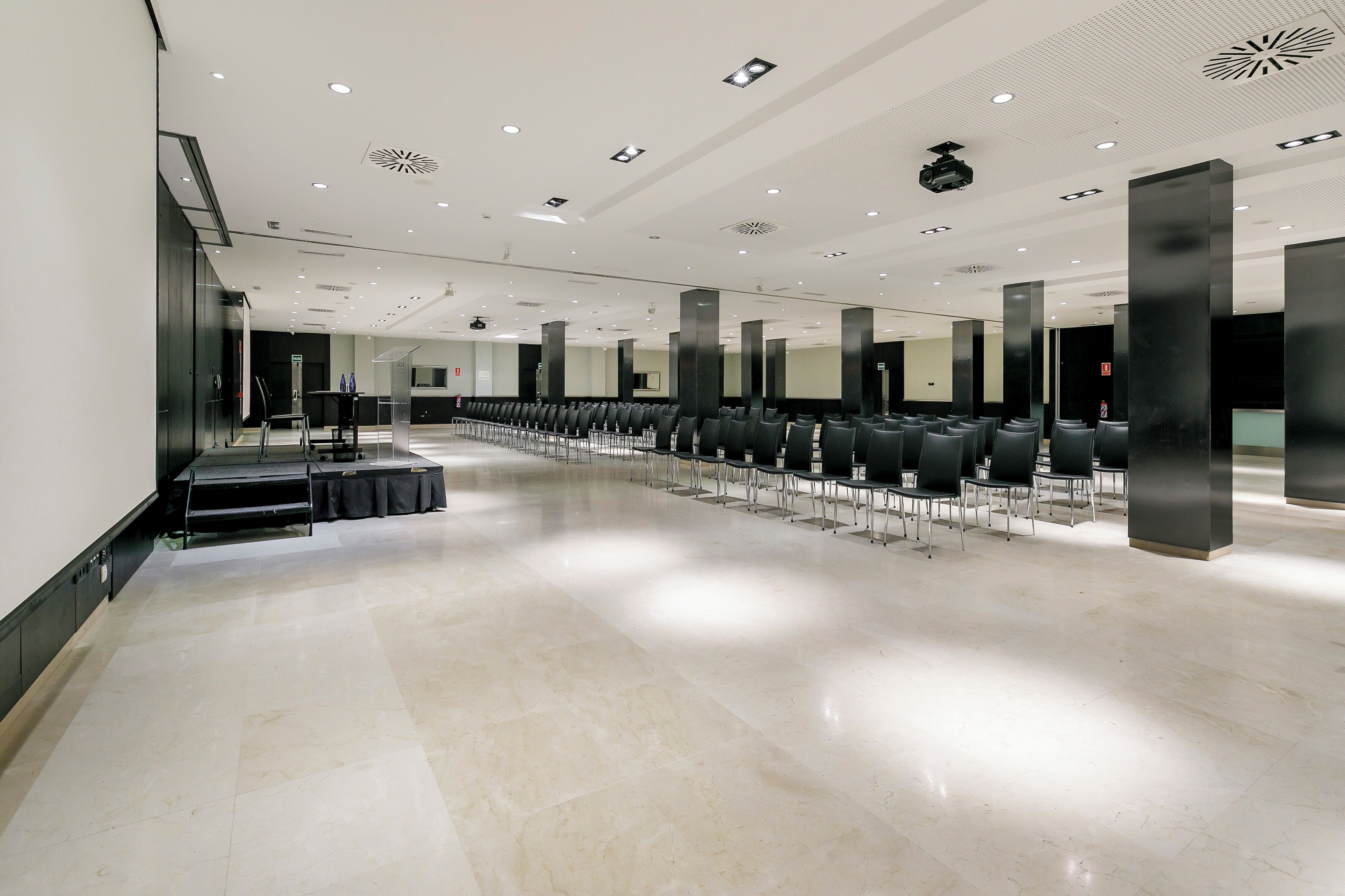 Een ruime hotellobby met rijen zwarte stoelen, een receptiebalie en een grote spiegelwand.