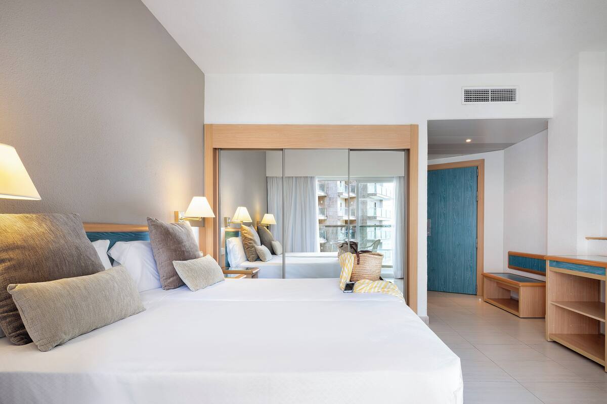 Habitación de hotel con una cama grande, lámparas de noche, un espejo, una ventana con vistas a edificios y un banco de madera.