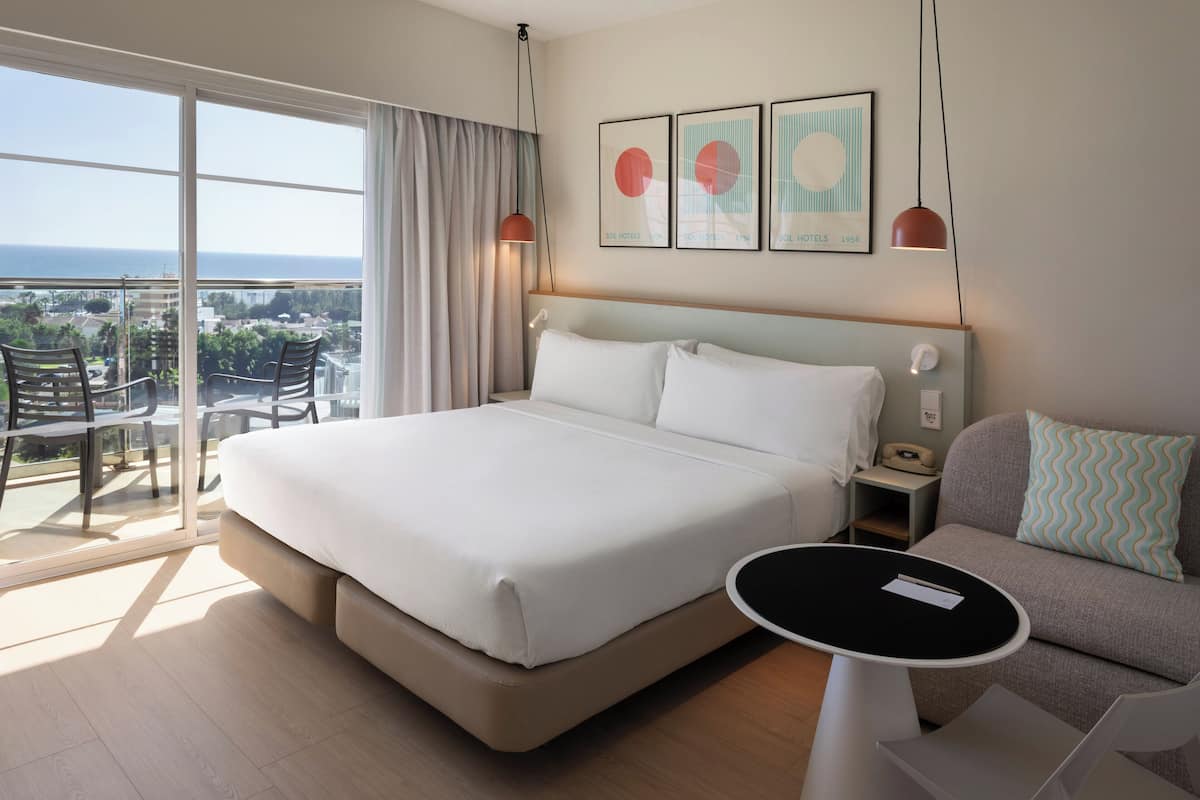 Habitación de hotel con una cama grande, un sofá, una mesita y un balcón con vistas a la ciudad.