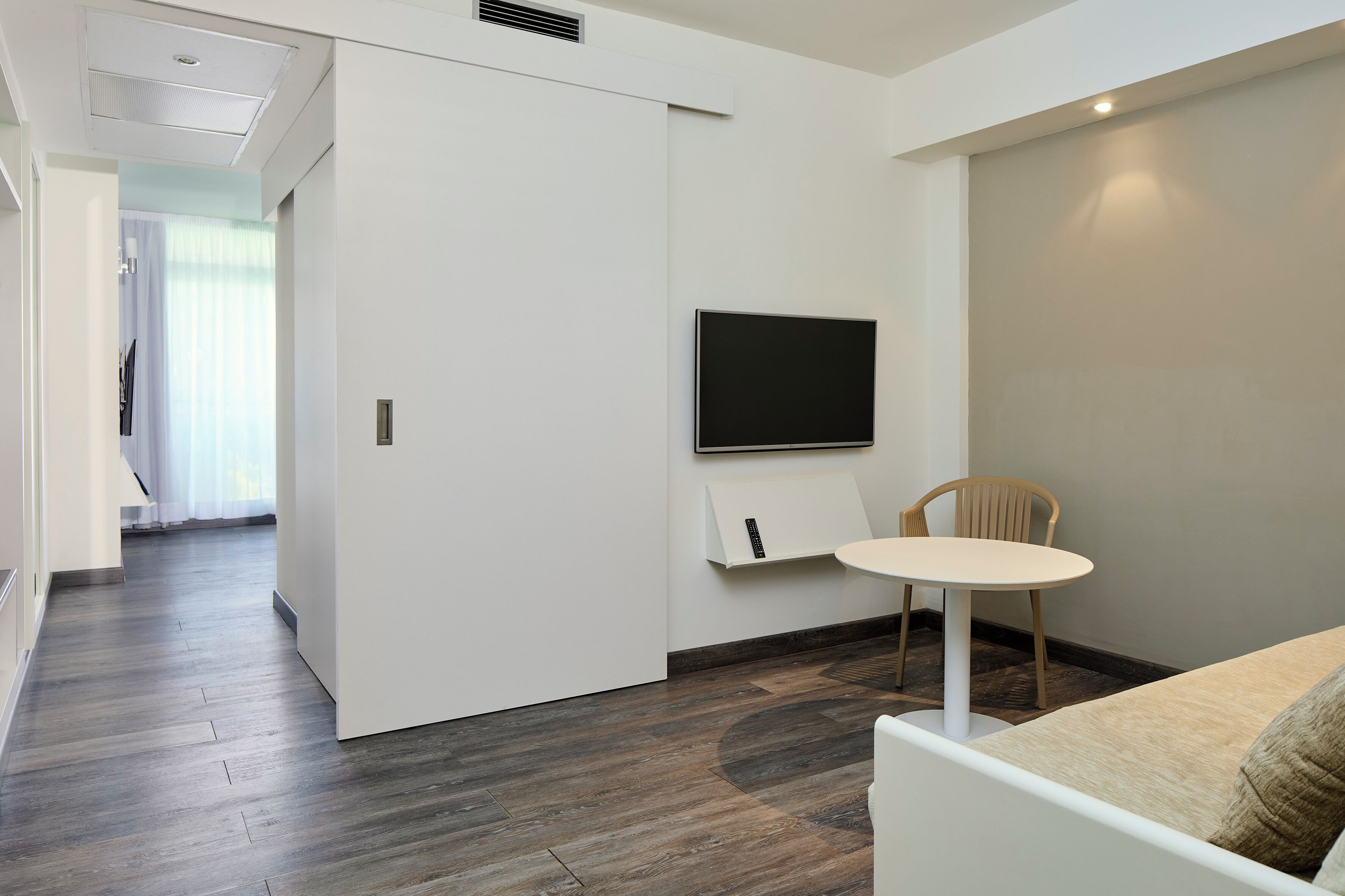 Una habitación de hotel moderna con piso de madera, un sofá blanco, una pequeña mesa redonda, un televisor montado en la pared y una puerta que da a otra habitación.