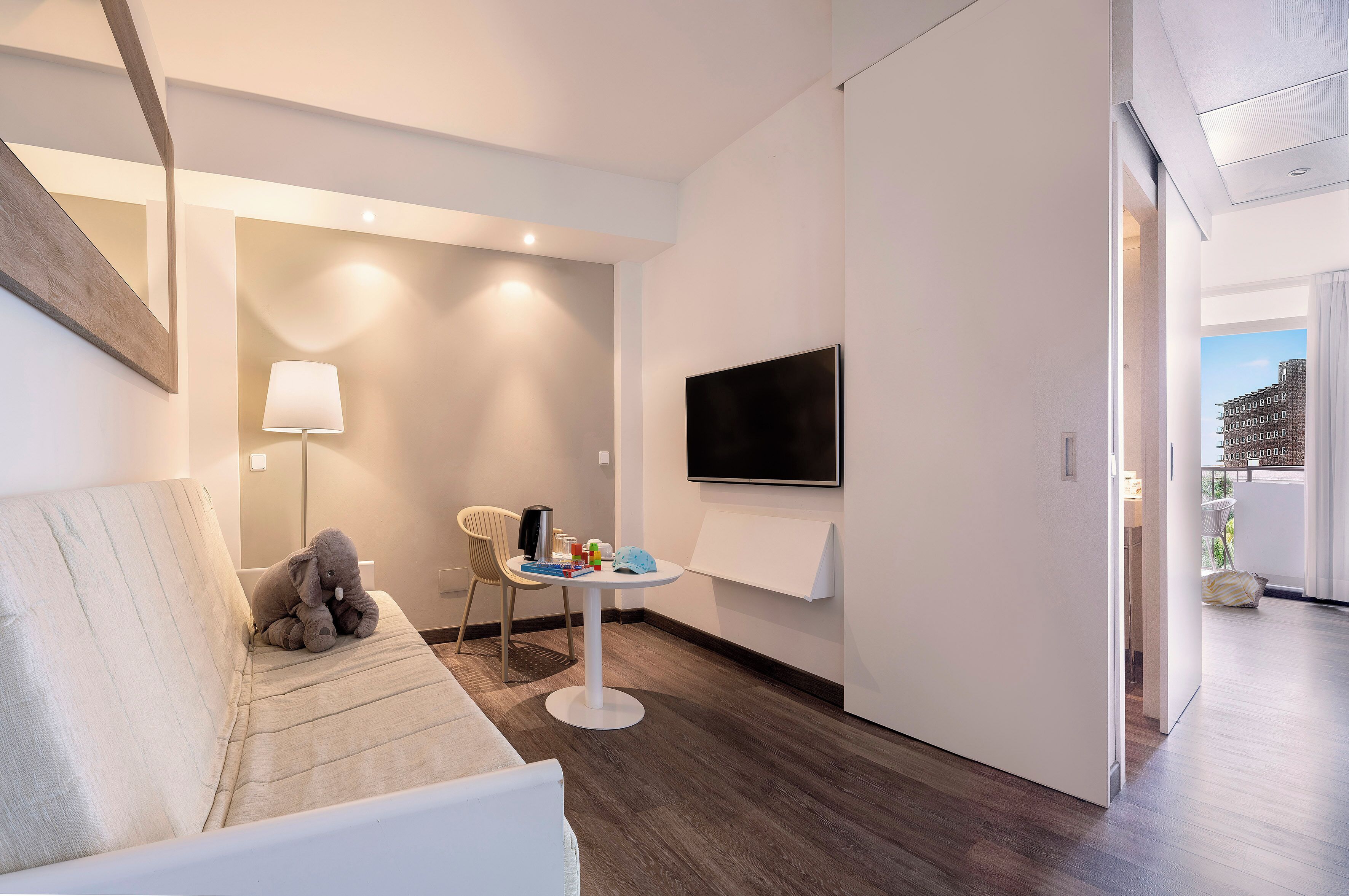 Habitació d'hotel moderna amb un sofà, una taula petita amb objectes, una làmpada, un televisor a la paret i vista cap a l'exterior.