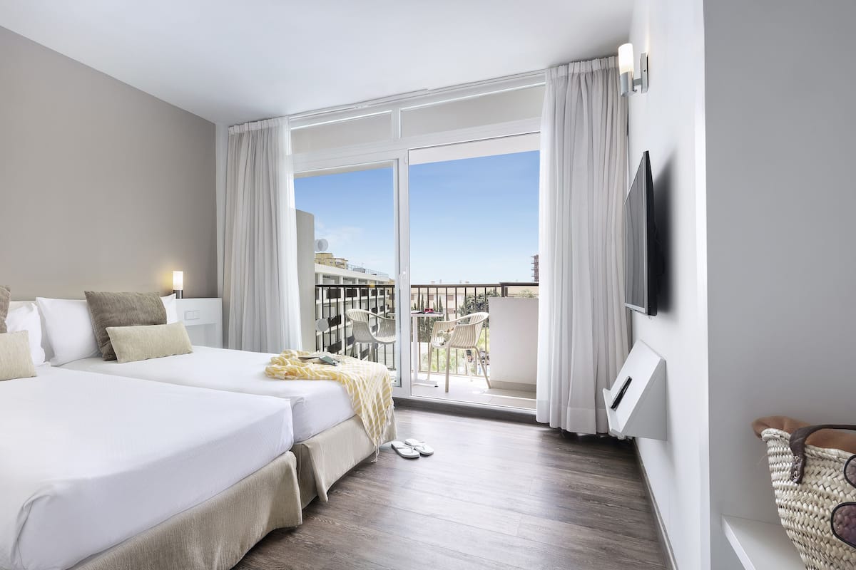 Habitación de hotel con dos camas, un balcón con vistas, un televisor de pantalla plana y una pequeña mesa con sillas.