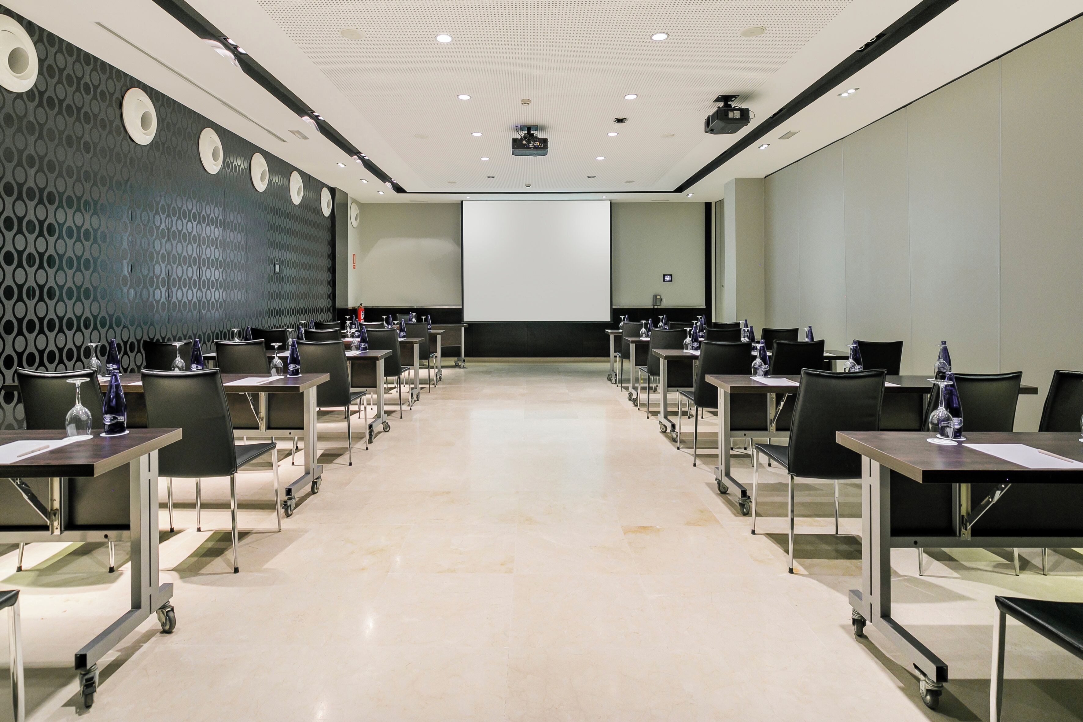 A conference room with a large screen, rows of tables with chairs, and a patterned wall.