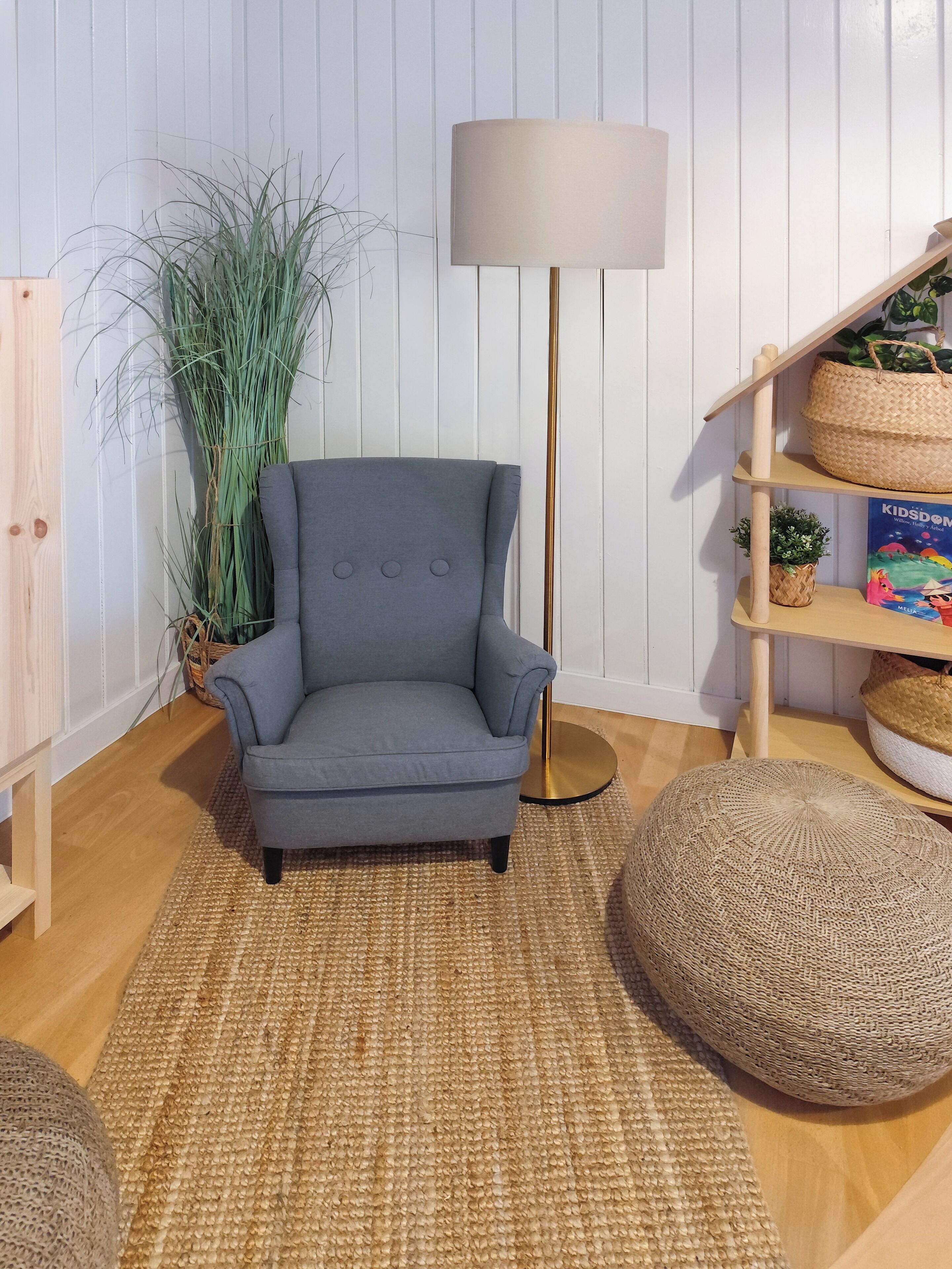 A room with a grey armchair, a floor lamp, a woven rug, a round pouf, and a bookshelf with a book and a woven basket.