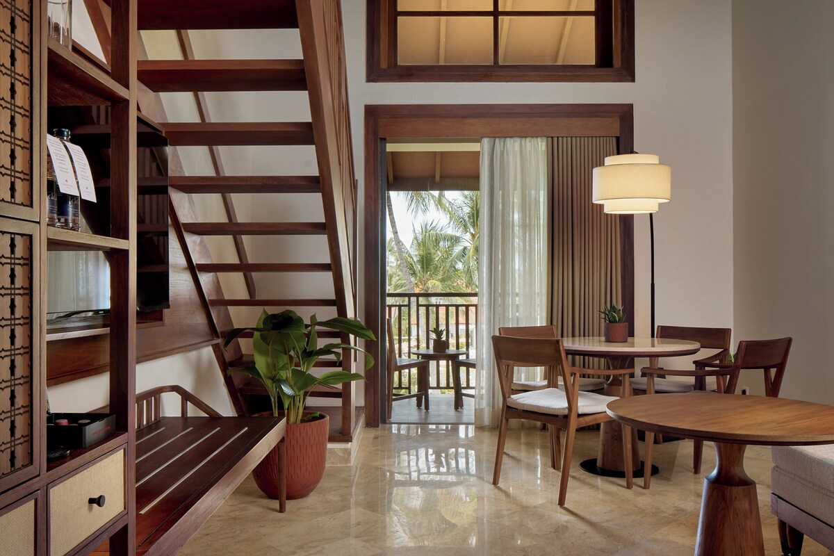 A wooden staircase, a dining area with chairs and a table, a potted plant, and a view of palm trees through a window.