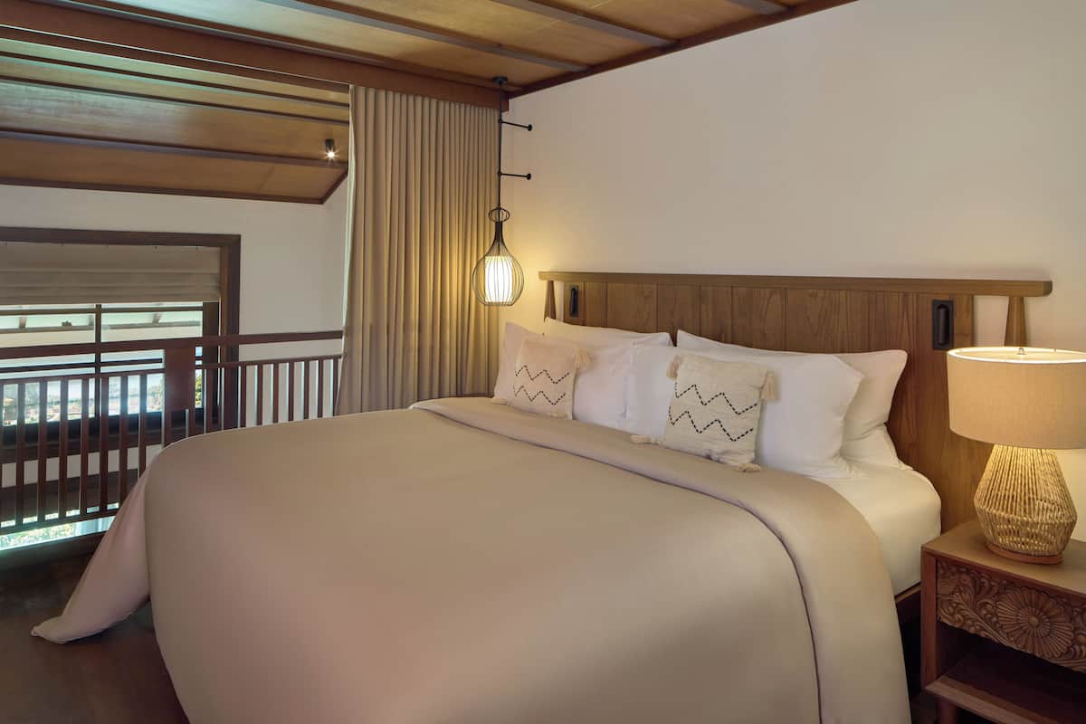 Ein Hotelzimmer mit einem großen Bett, einem Nachttisch mit Lampe, einem Fenster mit Vorhängen und einer Holzboden.