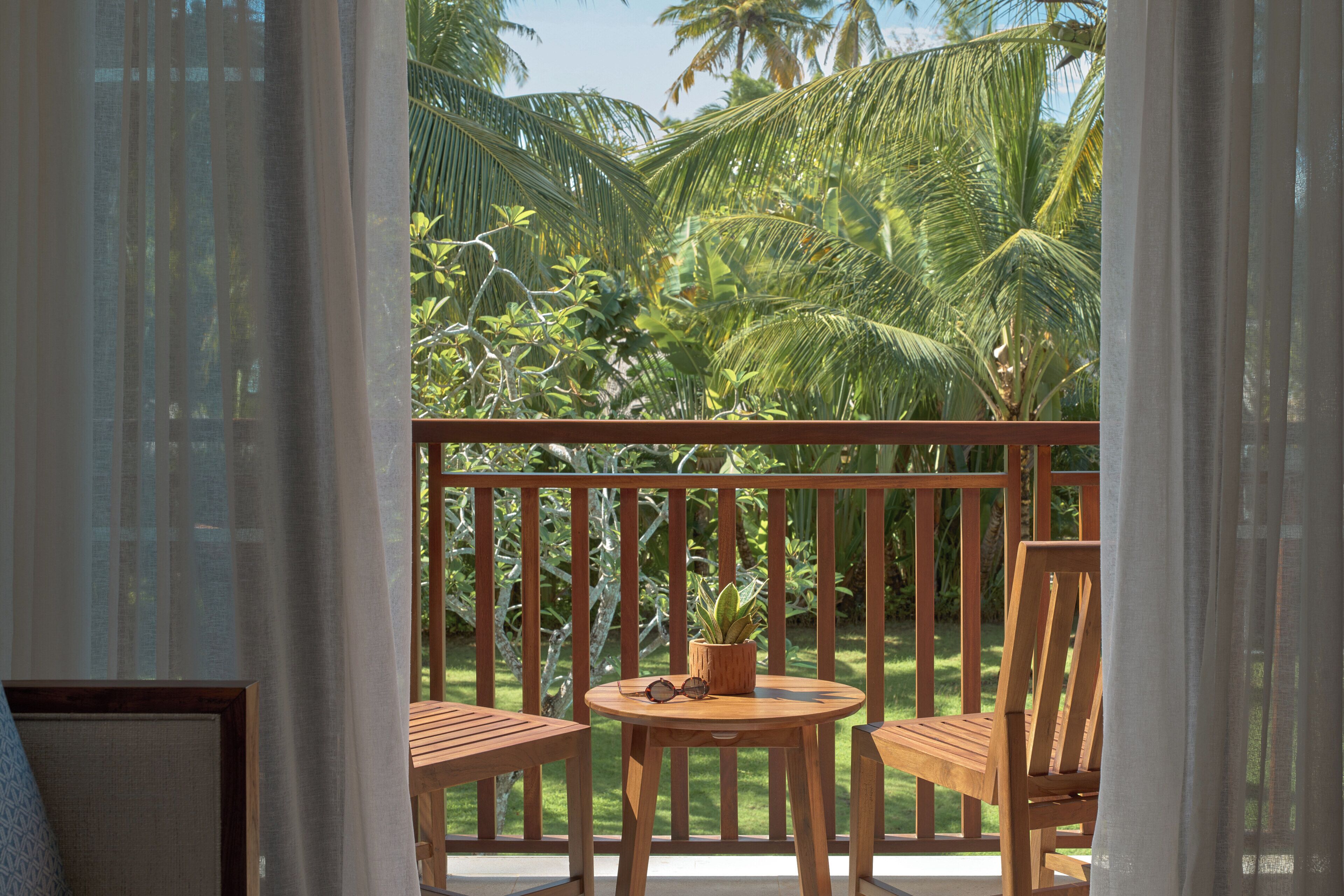 A wooden balcony with a table and chairs, a potted plant, and a view of tropical foliage.