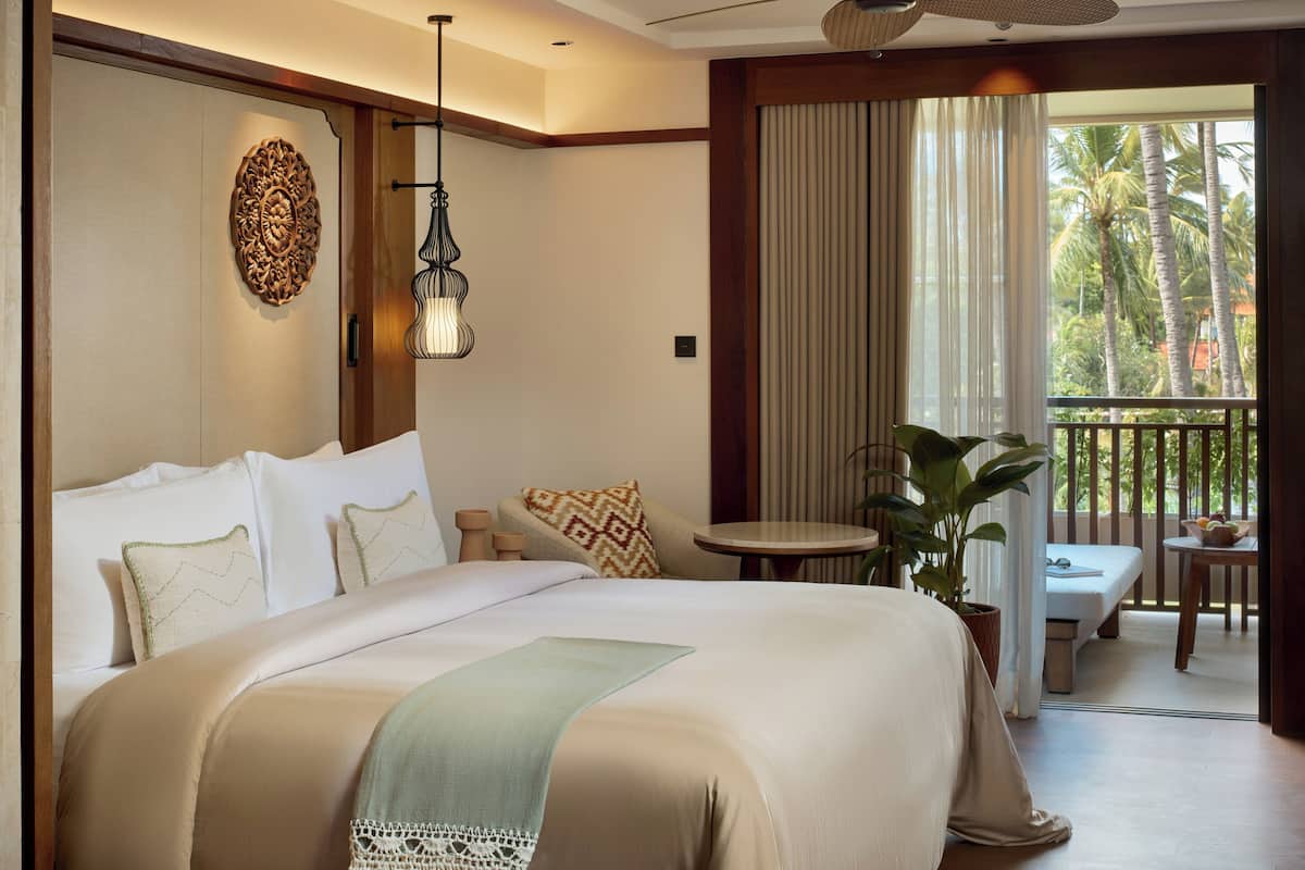 Ein Hotelzimmer mit einem großen Bett, einem kleinen Tisch, einer Zimmerpflanze und einem Balkon mit Blick auf Palmen.