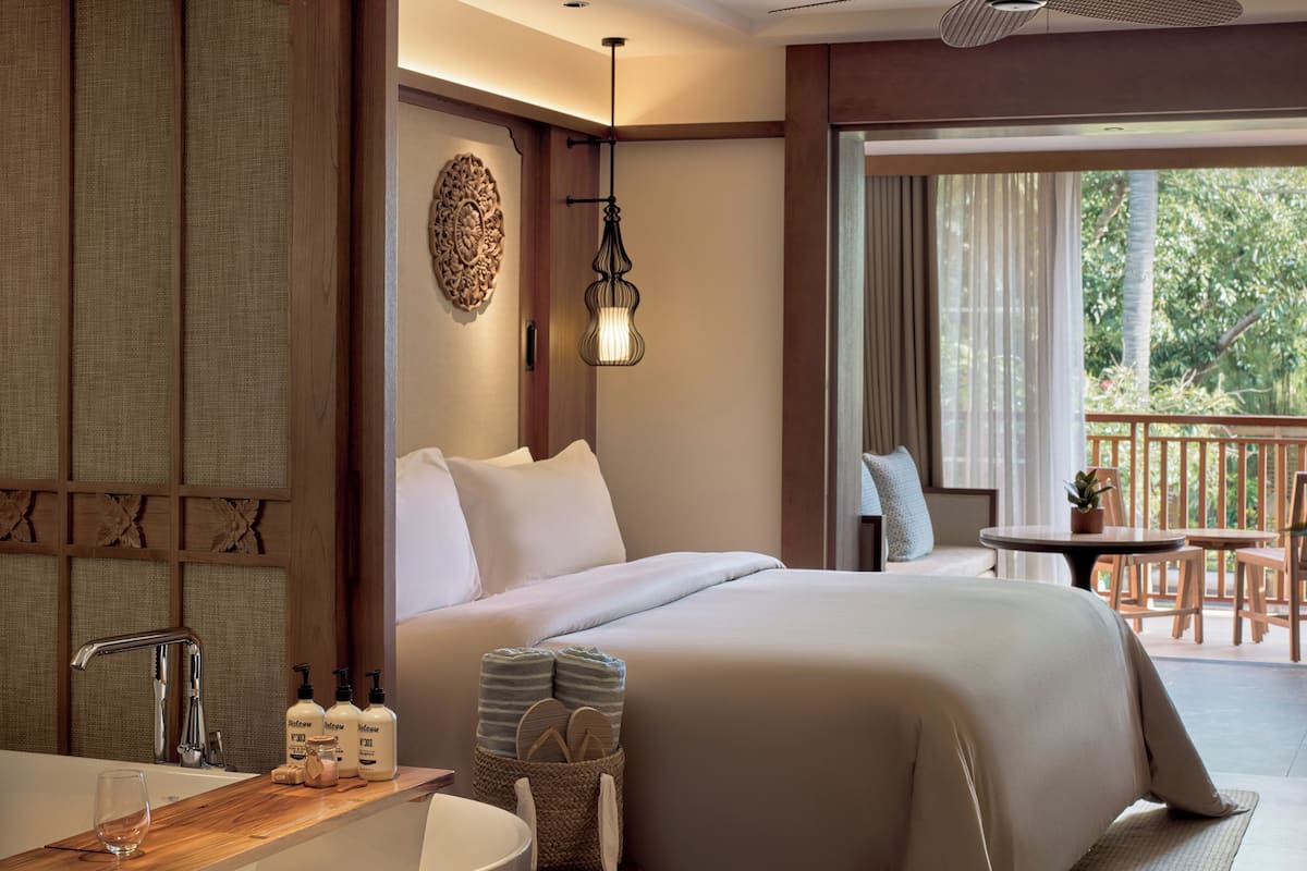 Ein Hotelzimmer mit einem großen Bett, einem Badezimmer mit Badewanne, einem Balkon mit Tisch und Stühlen und Blick auf grüne Vegetation.