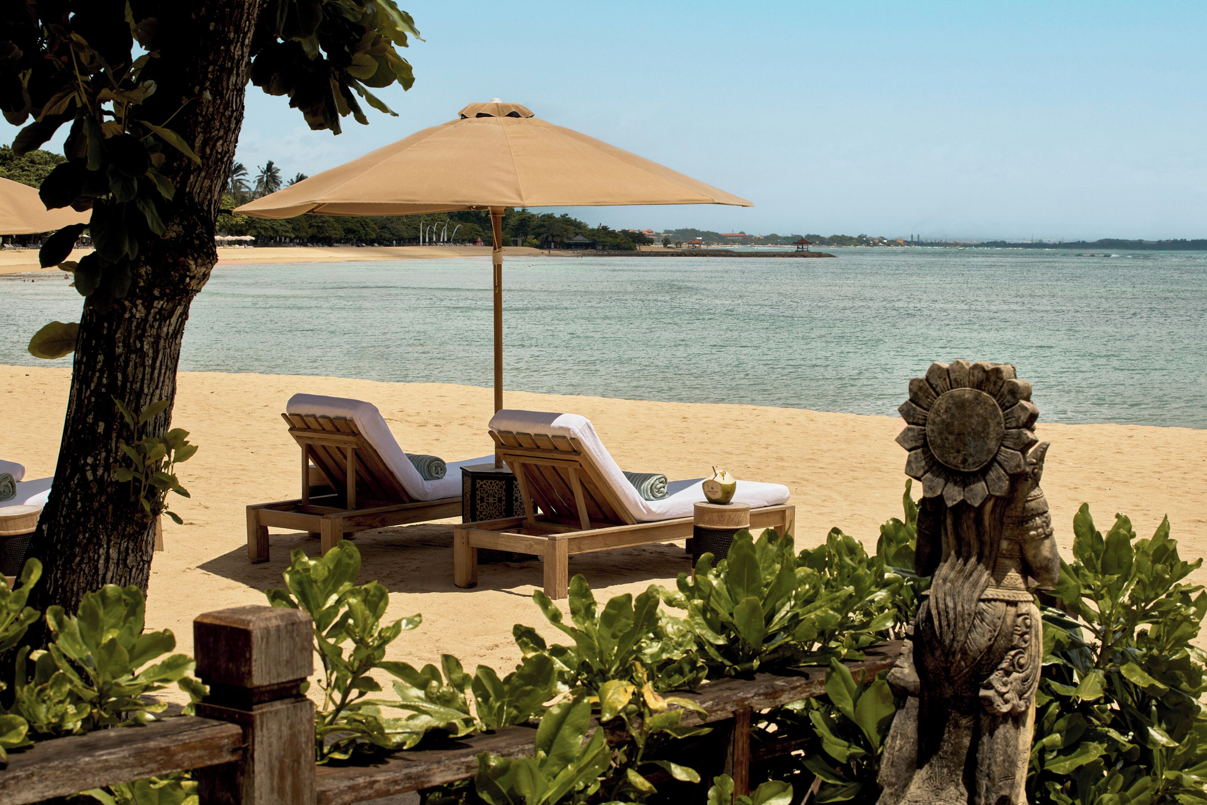 Un área de descanso junto a la playa con tumbonas y un gran parasol, rodeada de vegetación y una escultura de piedra decorativa.