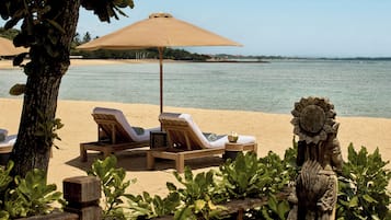 A beachside lounge area with sun loungers and a large umbrella, surrounded by greenery and a decorative stone sculpture.