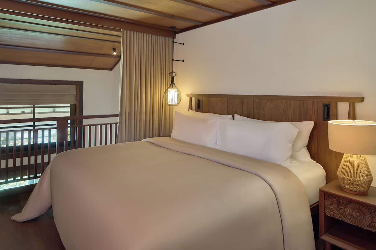 Ein Hotelzimmer mit einem großen Bett, einem Nachttisch mit Lampe, einem Fenster mit Vorhängen und einer Holzbalkendecke.