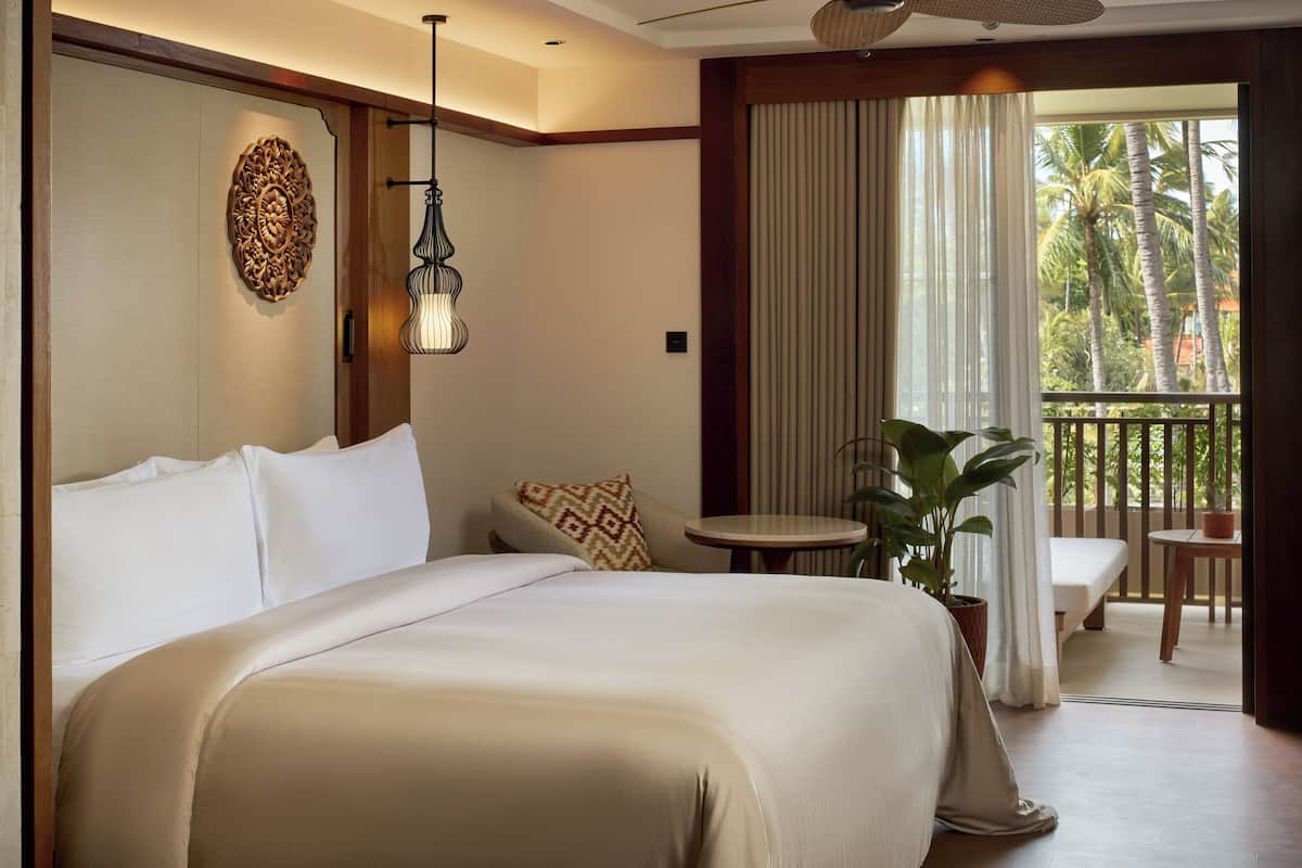 Ein Hotelzimmer mit einem großen Bett, einem kleinen Tisch, einer Topfpflanze und einem Balkon mit Blick auf Palmen.