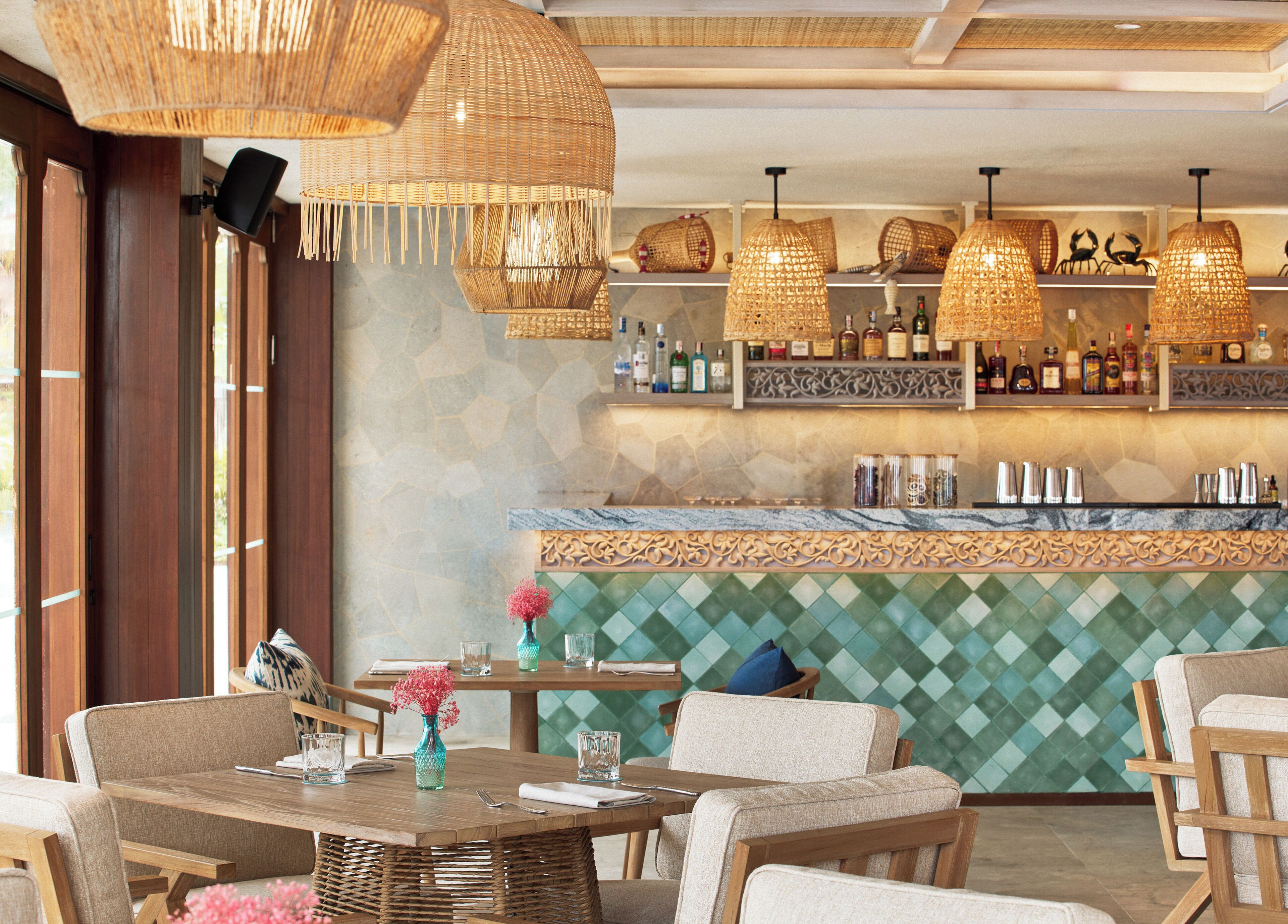 Un restaurante con lámparas colgantes de mimbre, una zona de bar con botellas y una pared verde con diseño de diamantes.