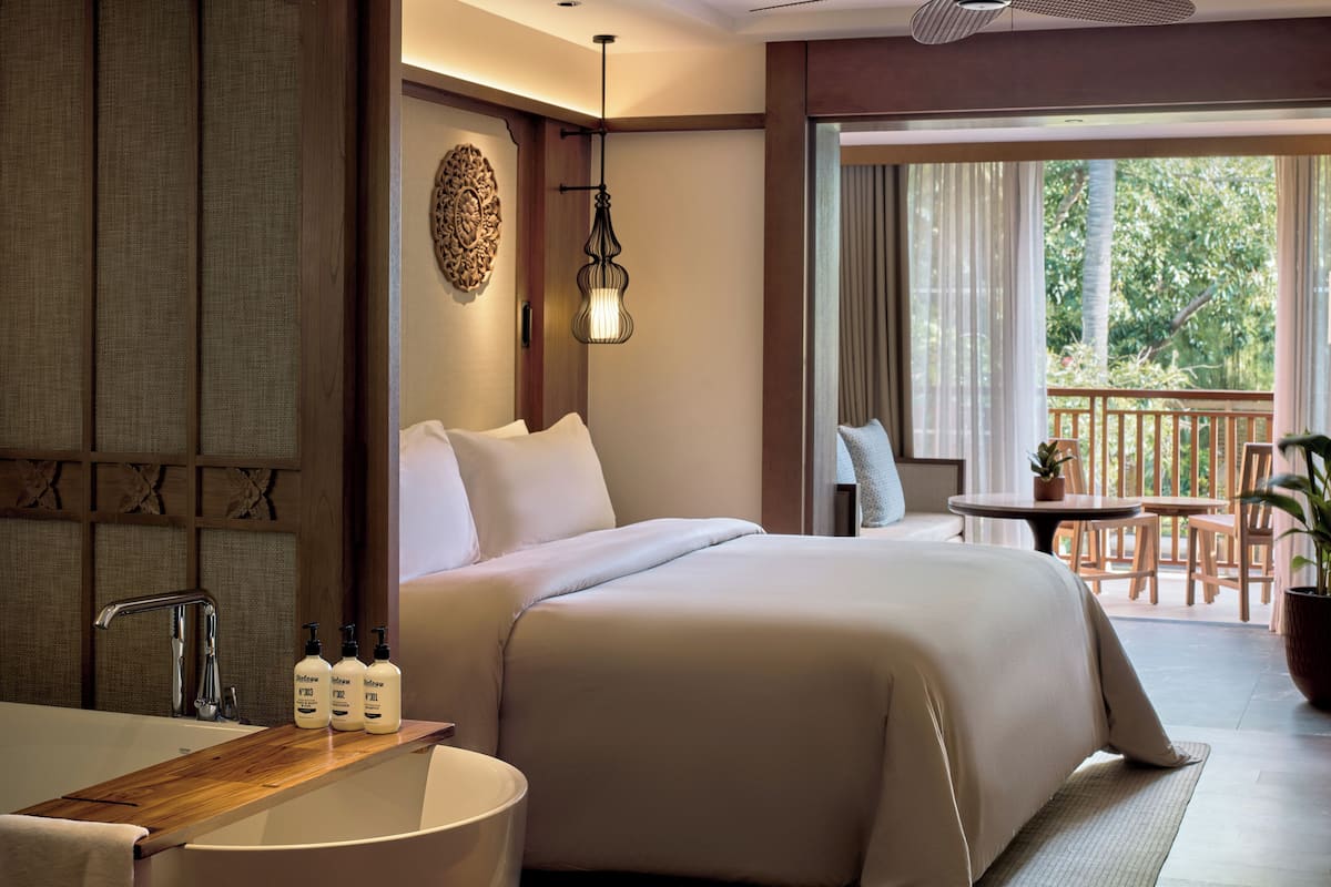Ein Hotelzimmer mit einem großen Bett, einem Holz-Waschtisch mit Badewanne, einem kleinen Tisch mit Stühlen und einem Balkon mit Blick auf Bäume.
