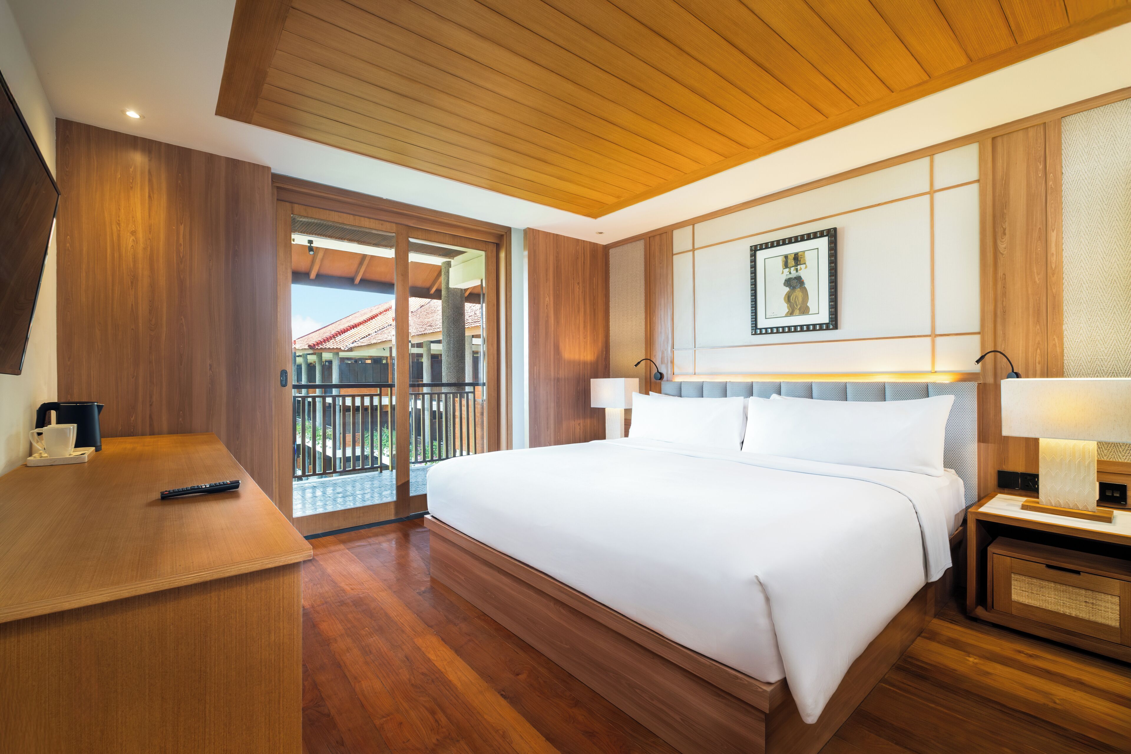 A hotel room with a wooden ceiling, a large bed with white linens, a desk with a remote control, and a balcony with a view of a red-roofed building.