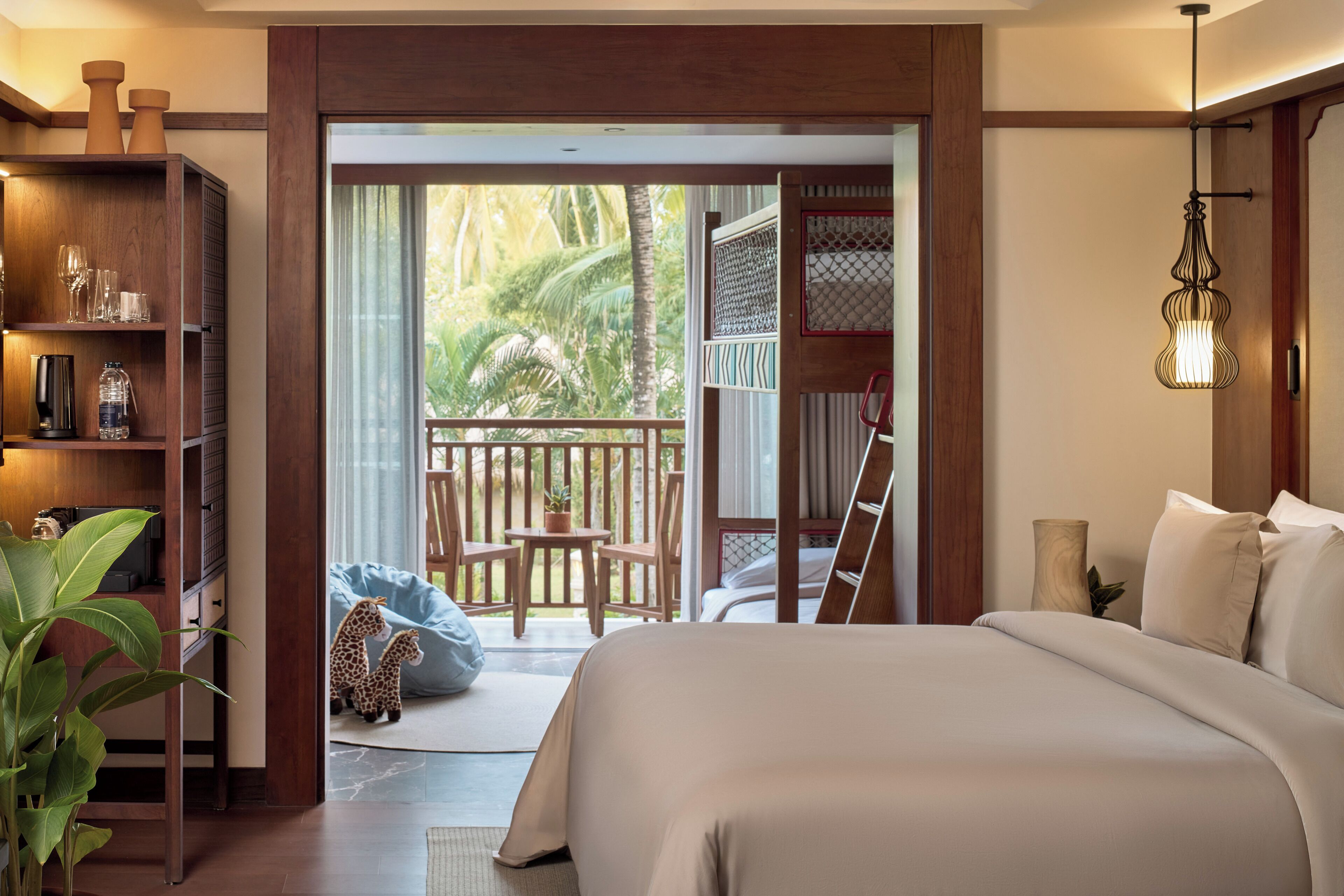 A bedroom with a large bed, a wooden bookshelf, a desk with a chair, and a balcony with a view of greenery.