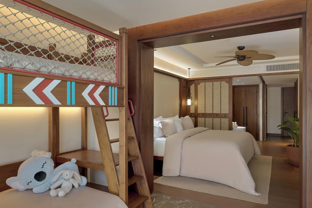 Ein Schlafzimmer mit Etagenbett, einer Himmellaune, einem Stofftier, einem Bett mit weißer Bettwäsche, einem Deckenventilator und Blick in einen angrenzenden Raum.
