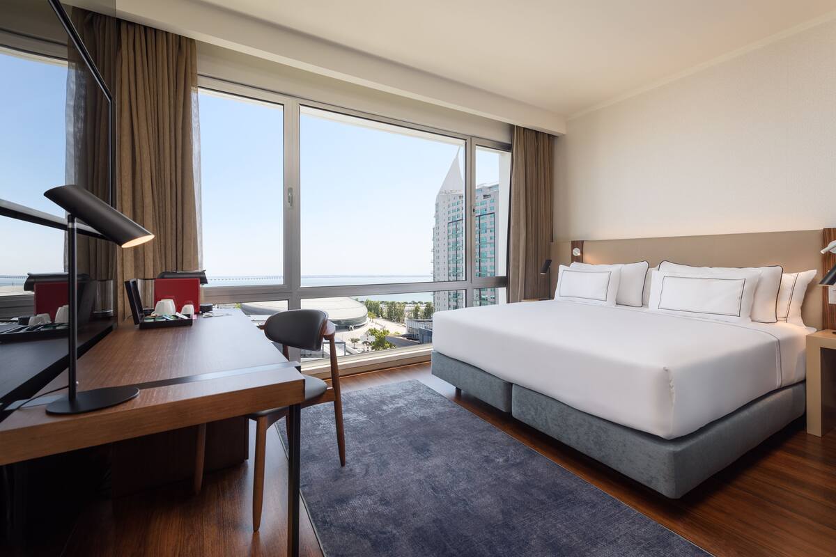 Una habitación de hotel moderna con una cama grande, un escritorio, una silla y vistas a la ciudad a través de amplios ventanales.