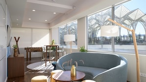 A modern living room with a grey sofa, round coffee table, and two armchairs. There is a wooden side table with a vase and a lamp. Large windows offer a view of a glass structure outside.