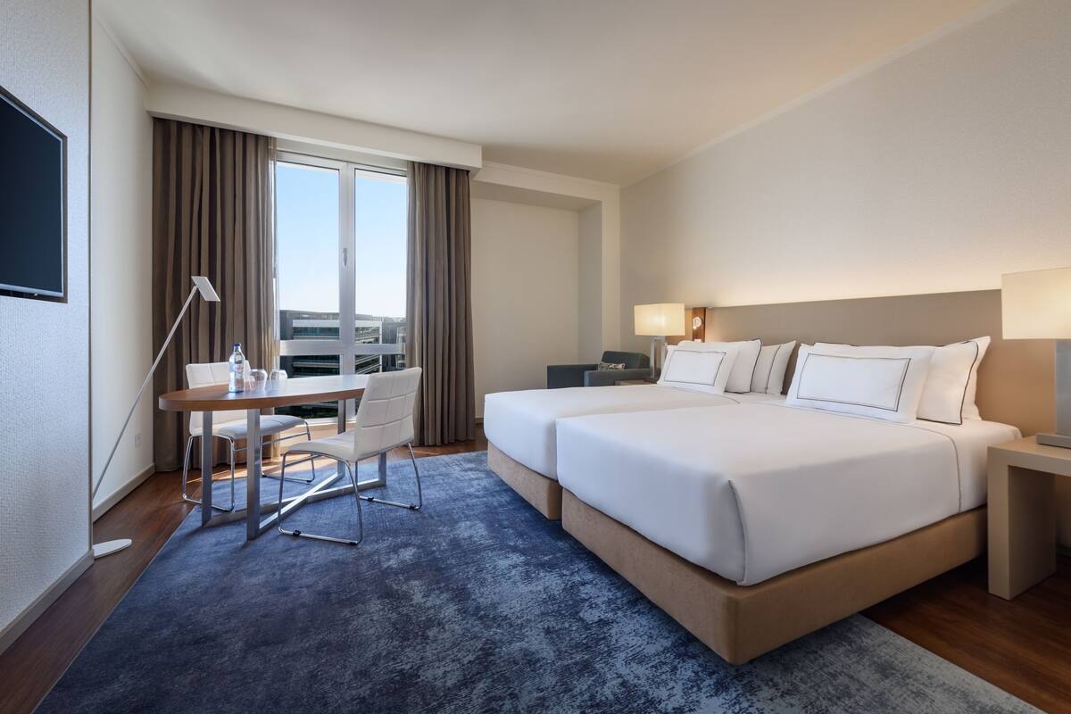 Habitación de hotel con una cama grande, un escritorio, una silla, un ventanal con cortinas y vistas a la ciudad.