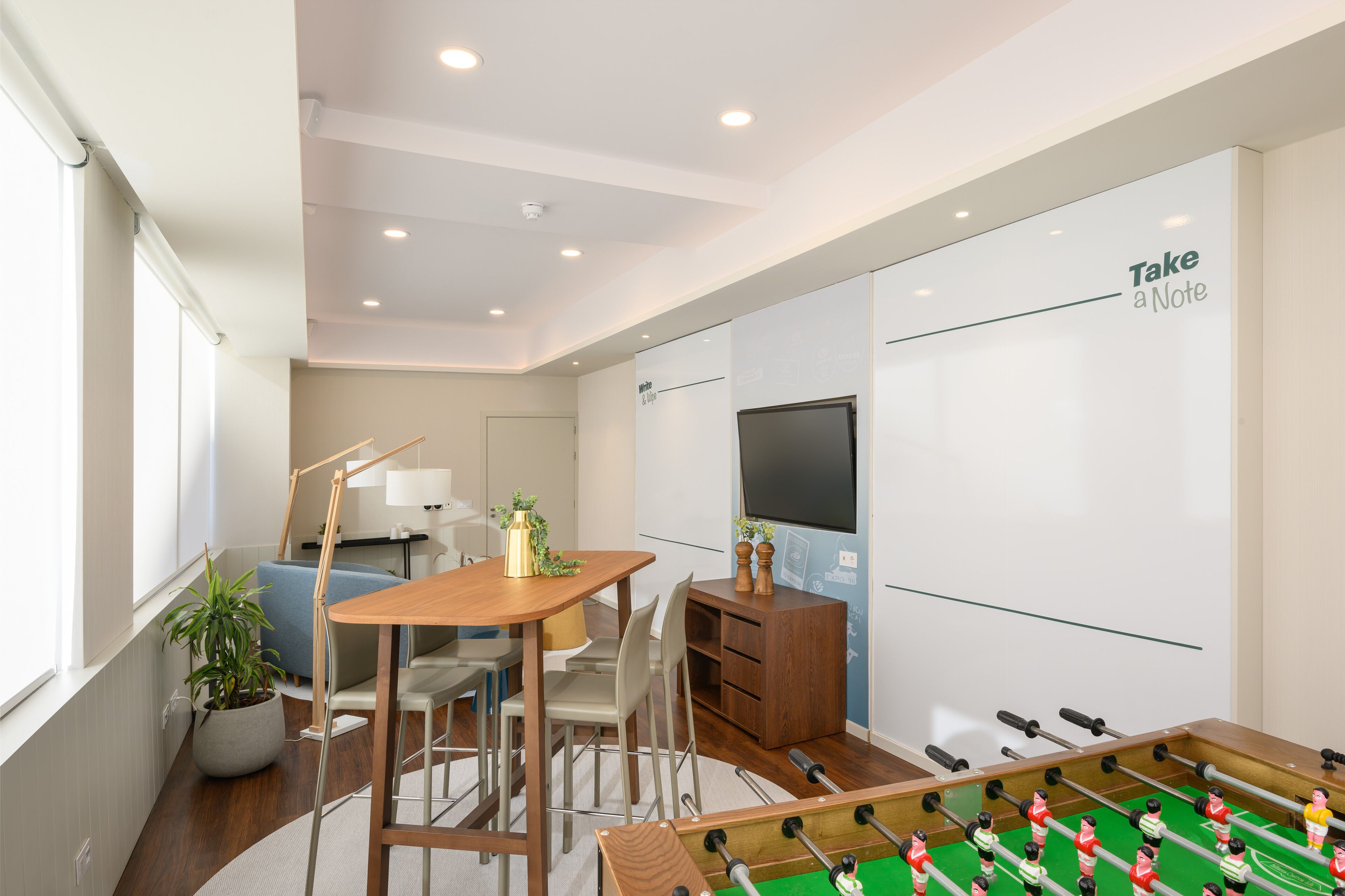 Una habitación moderna con mesa de futbolín, escritorio, sillas, planta en maceta y televisor en la pared.