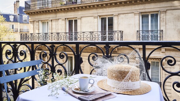 A balcony with a table set for two, a cup of coffee, a straw hat, and a decorative item.